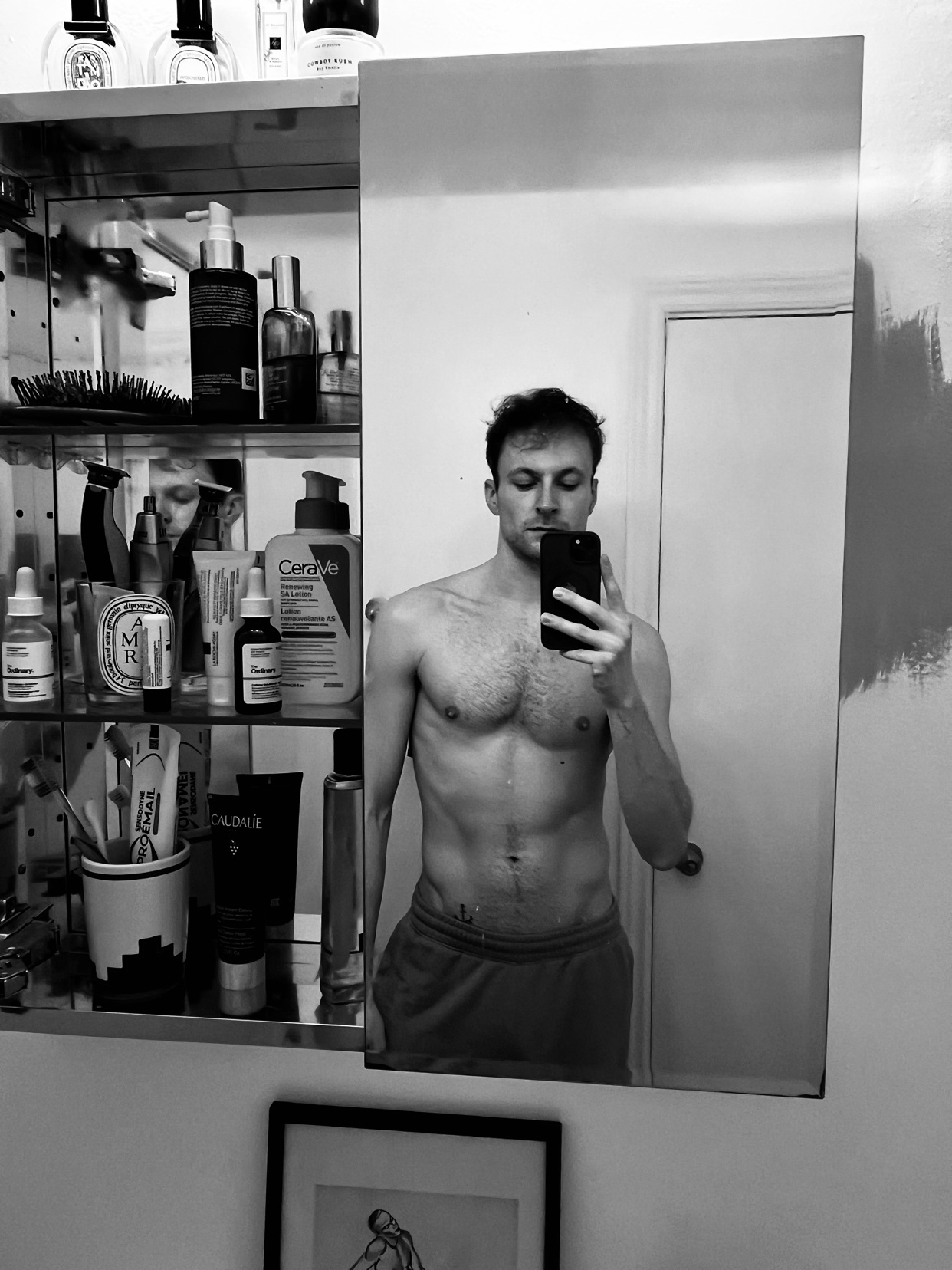A man takes a mirror selfie, shirtless, showcasing his physique. The background reveals a bathroom with various grooming products.