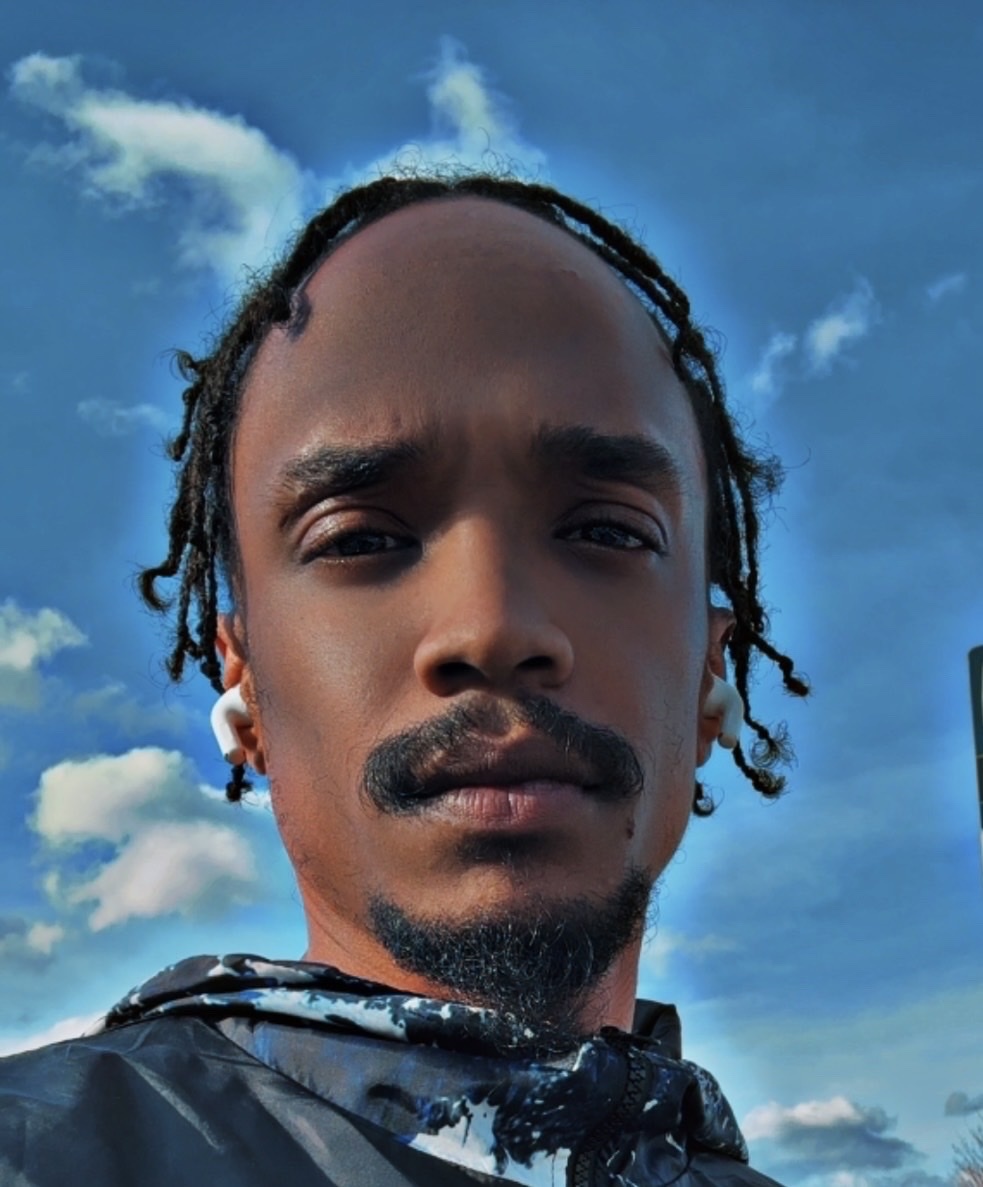 A close-up, low-angle shot of a man with dreadlocks, a mustache, and a goatee. He is wearing wireless earbuds and a jacket, with a blue sky and clouds in the background.