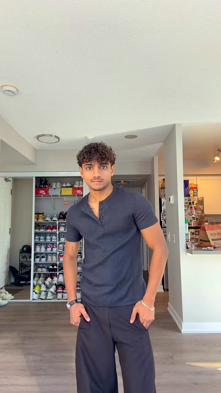 A young man with curly dark hair and a light beard poses indoors wearing a dark blue henley shirt and black wide-leg pants. He has his hands in his pockets and is looking towards the camera.
