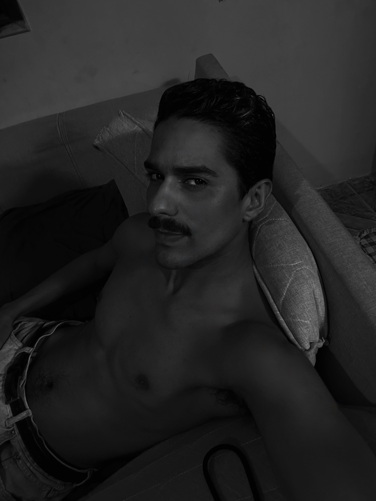 A black and white selfie of a man with a mustache, shirtless, lying on a couch.