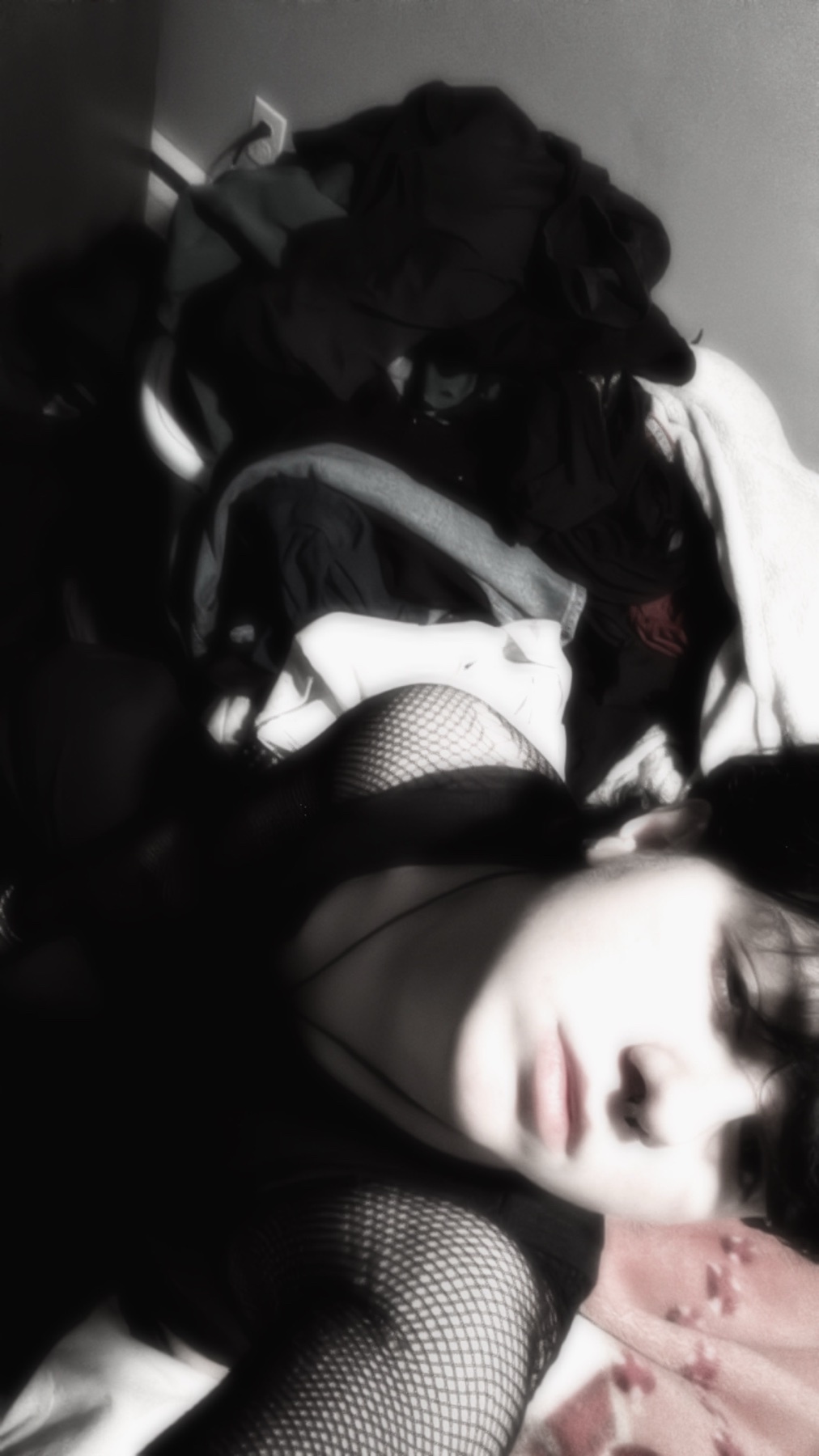 A woman with dark hair and fishnet clothing lies on a bed with a pile of clothes behind her. The lighting creates a dramatic shadow across her face.