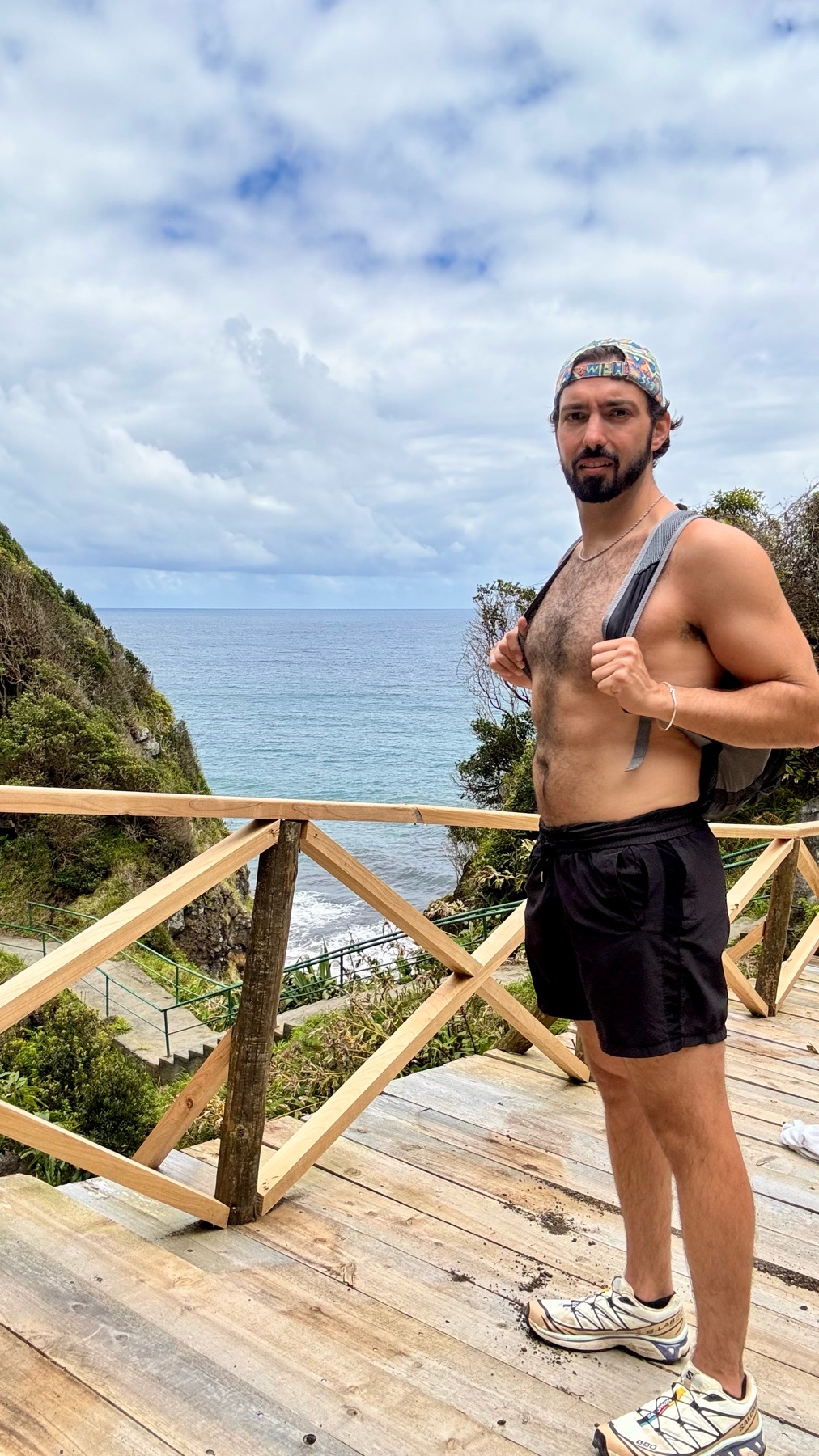 A shirtless man with a backpack and hat stands on a wooden walkway overlooking a coastal landscape with the ocean and cloudy sky in the background.
