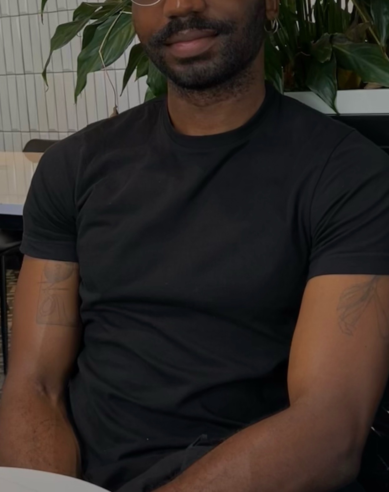 A man with a beard and glasses is wearing a black t-shirt. He has tattoos on his arms.