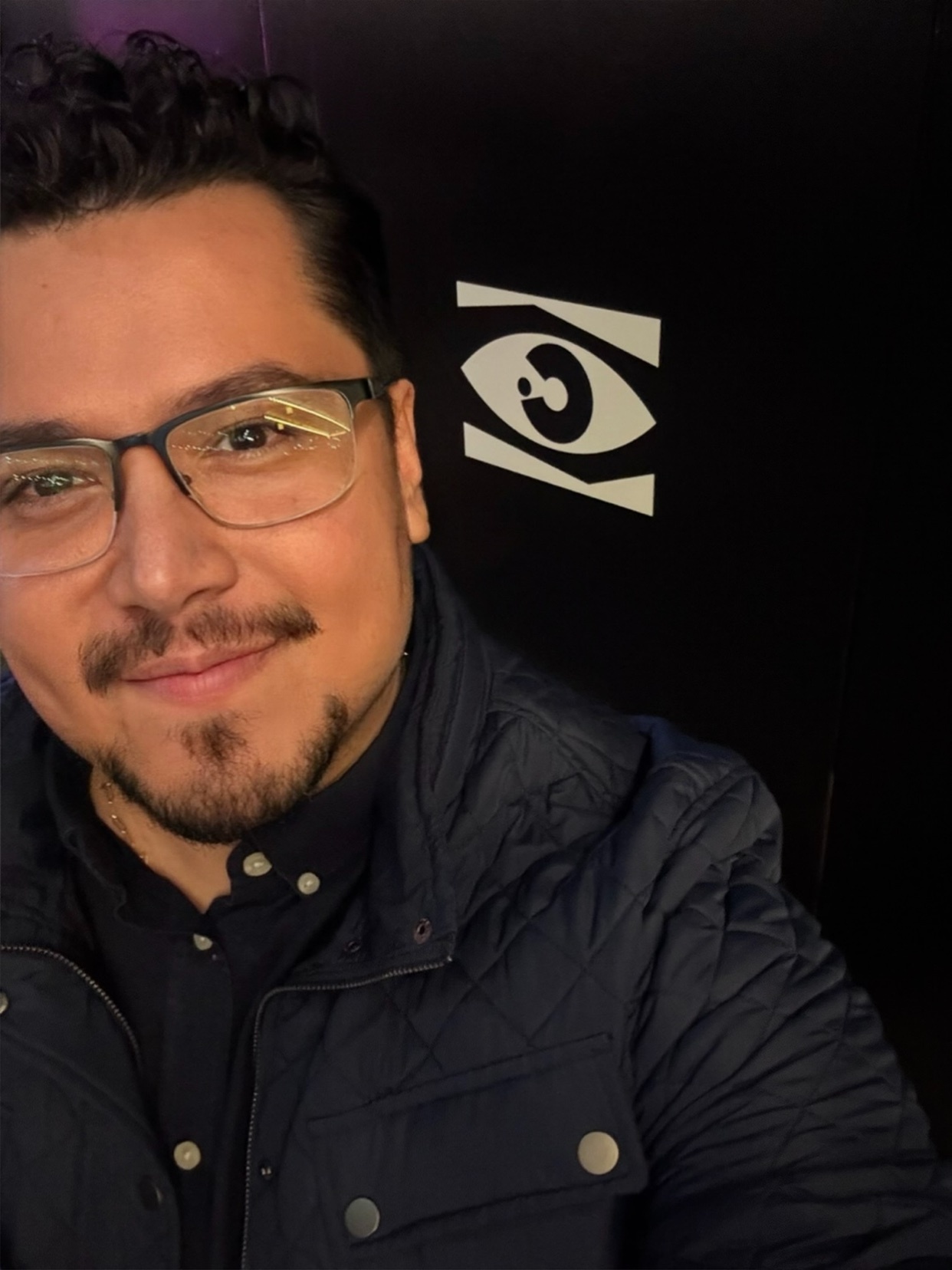 A man with glasses and a beard smiles at the camera. He is wearing a dark jacket and is in a club.