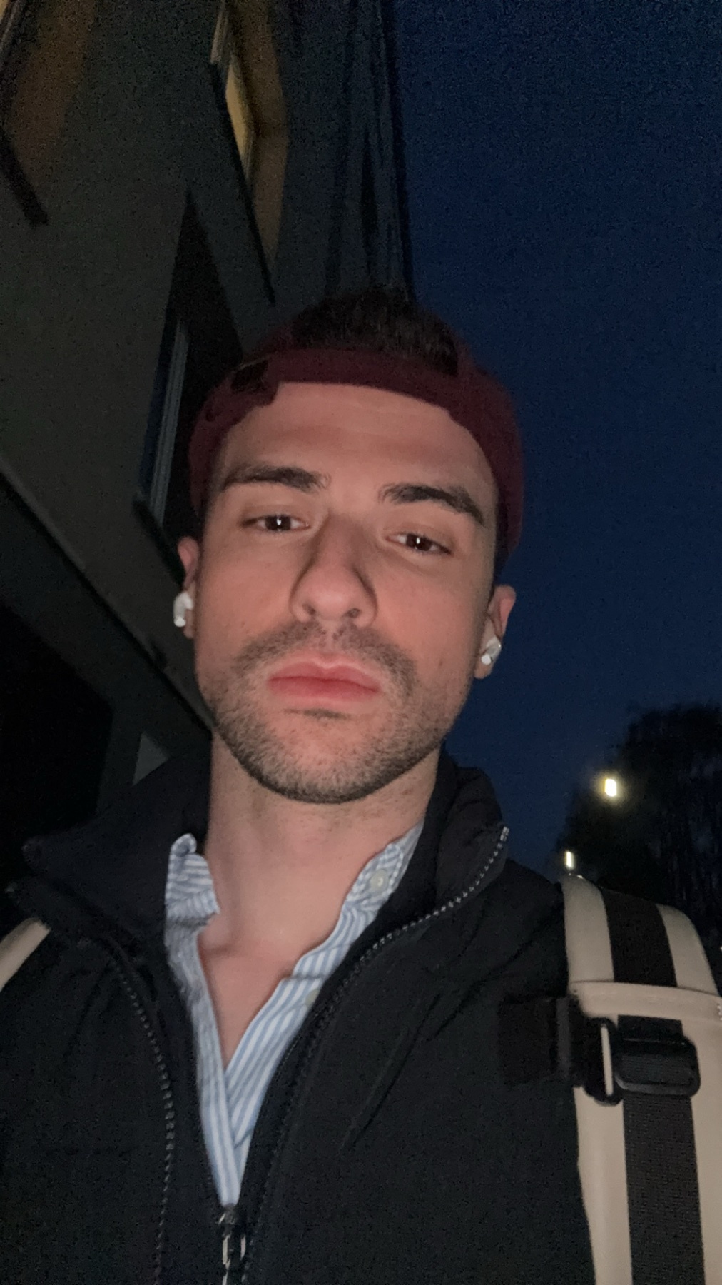 A man takes a selfie at night. He is wearing a cap and earbuds.