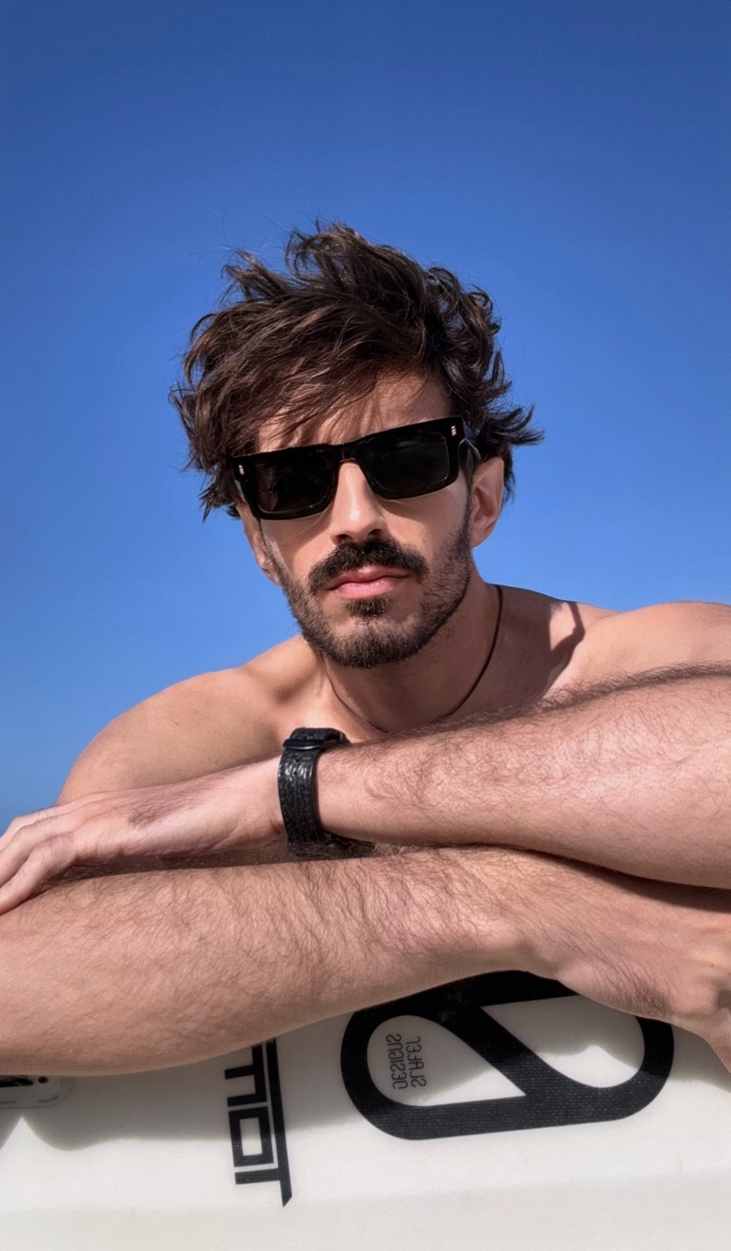 A man with dark hair and a beard poses shirtless, wearing sunglasses. He leans on a surfboard against a bright blue sky.