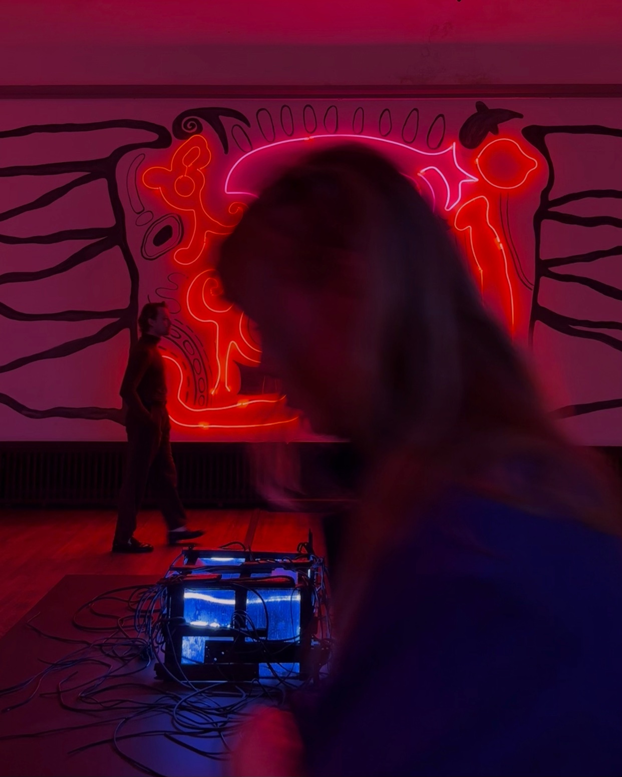 A blurry foreground of a woman's shoulder and head is in focus, with a man walking in the background past a large, vibrant neon art installation.