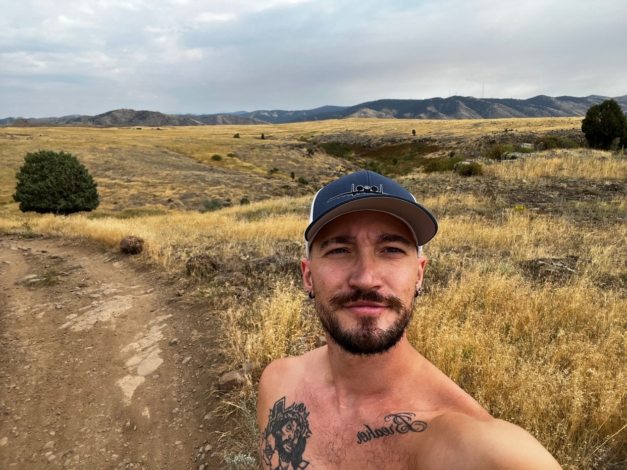 A man takes a selfie on a trail in a golden landscape. He is shirtless and wearing a cap.