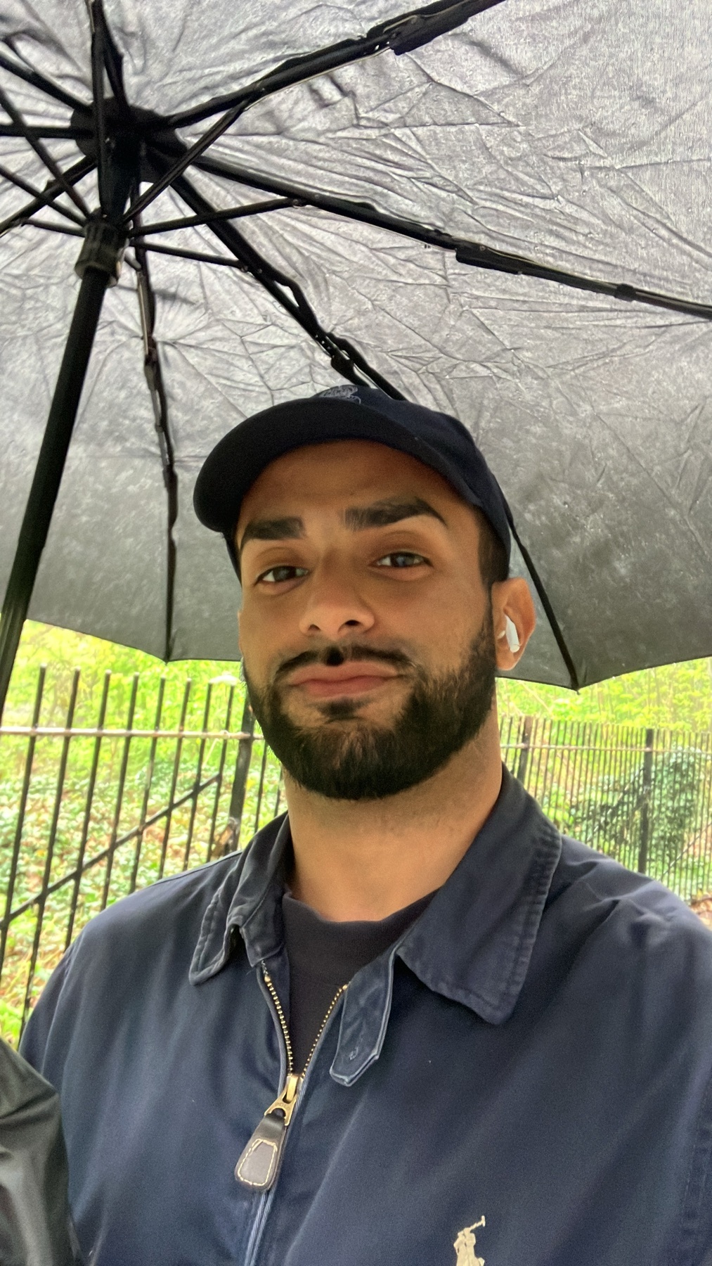 A man with a beard and stubble wears a baseball cap and a navy jacket under a black umbrella. He is looking at the camera with earbuds in his ears.