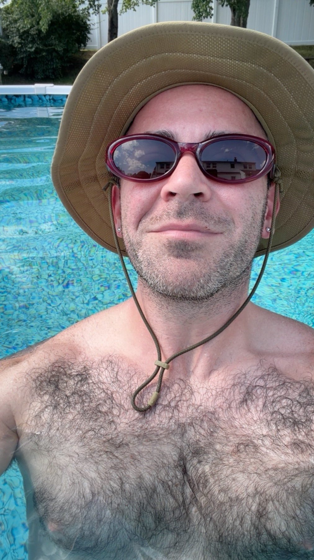 A man wearing a bucket hat and sunglasses smiles at the camera while in a swimming pool. He has a lot of chest hair and is enjoying pool time on the Fourth of July.