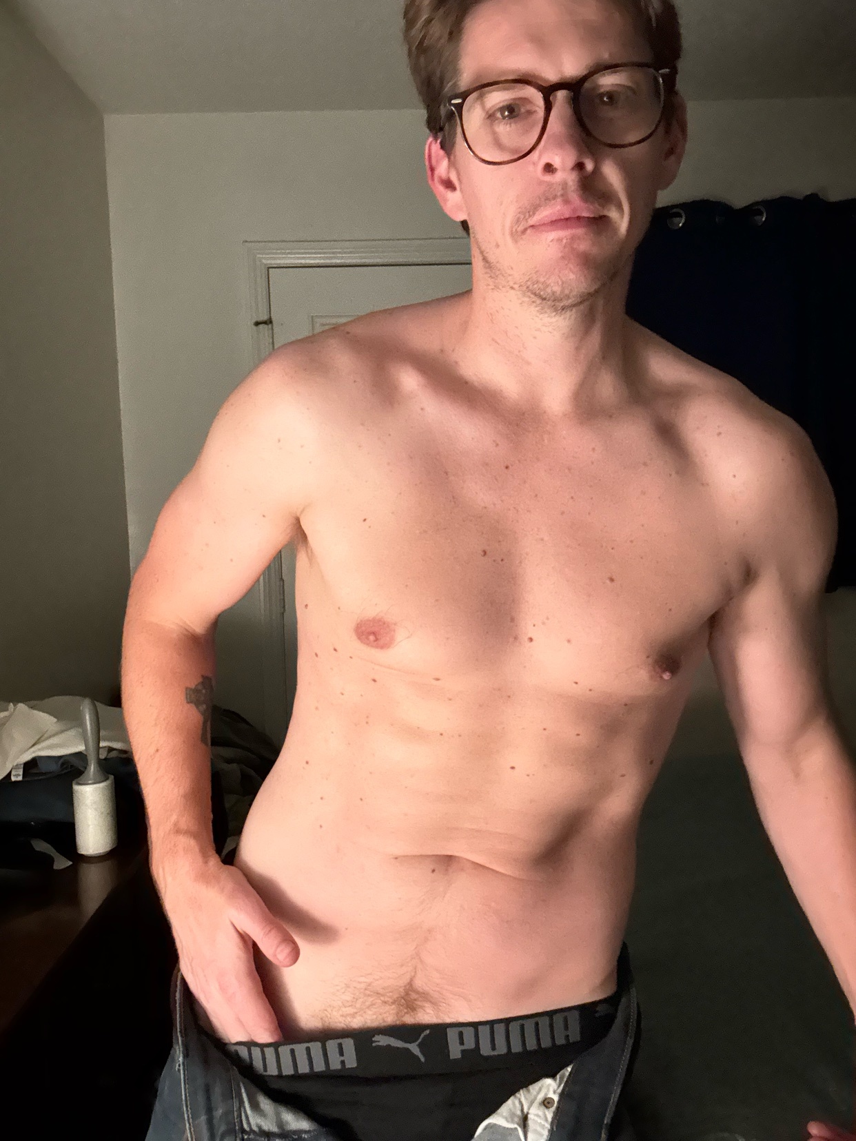 A man with glasses and a tattoo on his arm poses in his underwear and jeans.