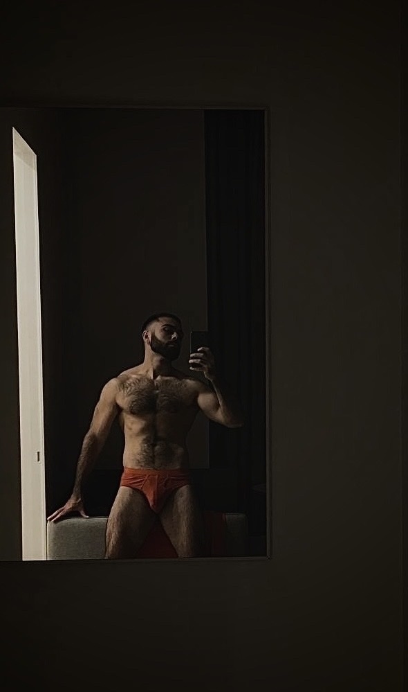 A man poses shirtless in front of a mirror, wearing red underwear. The photo highlights his muscular physique and chest hair.