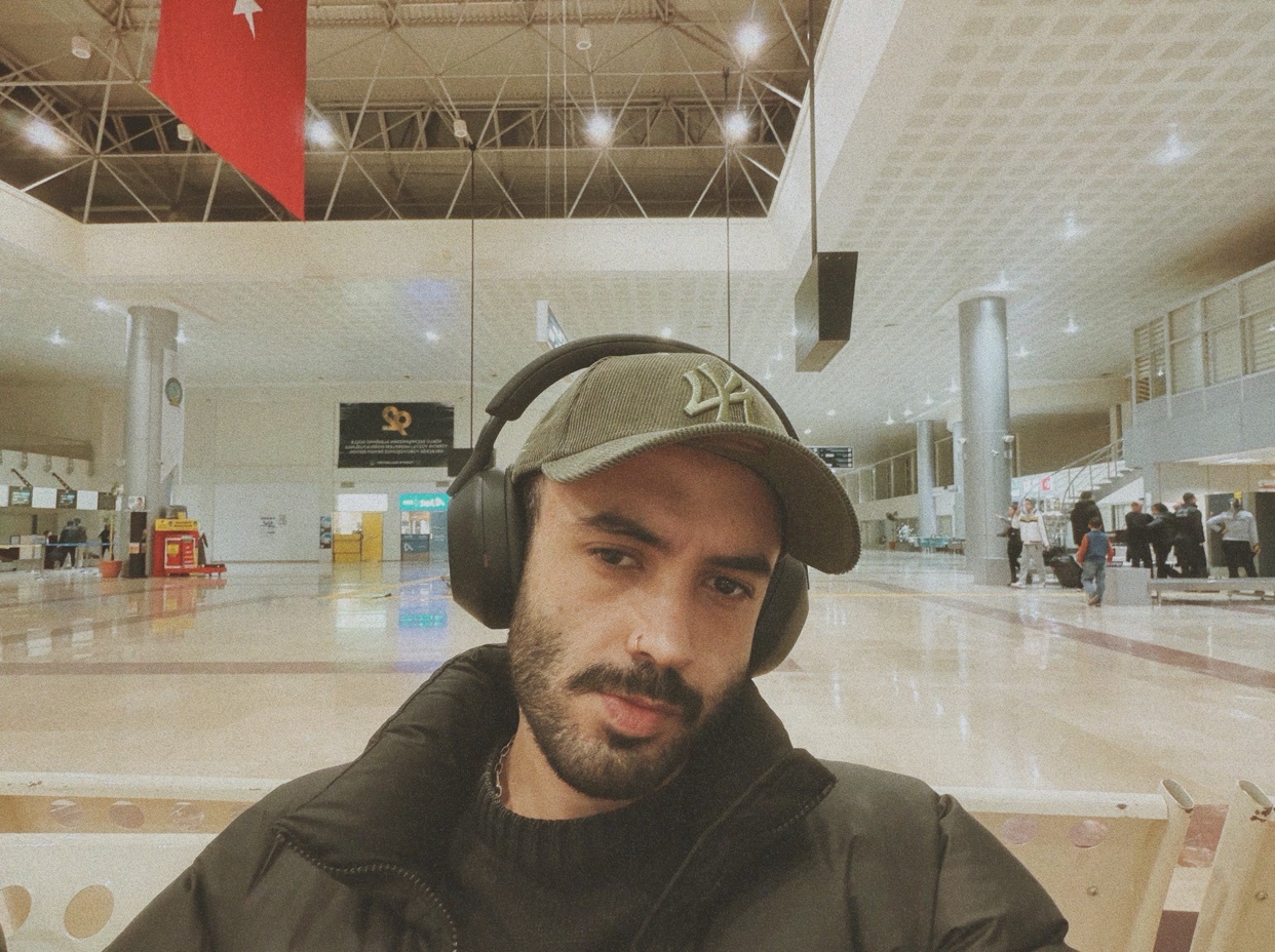 A man wearing a cap and headphones sits in an airport. The photo is taken indoors with a modern aesthetic.
