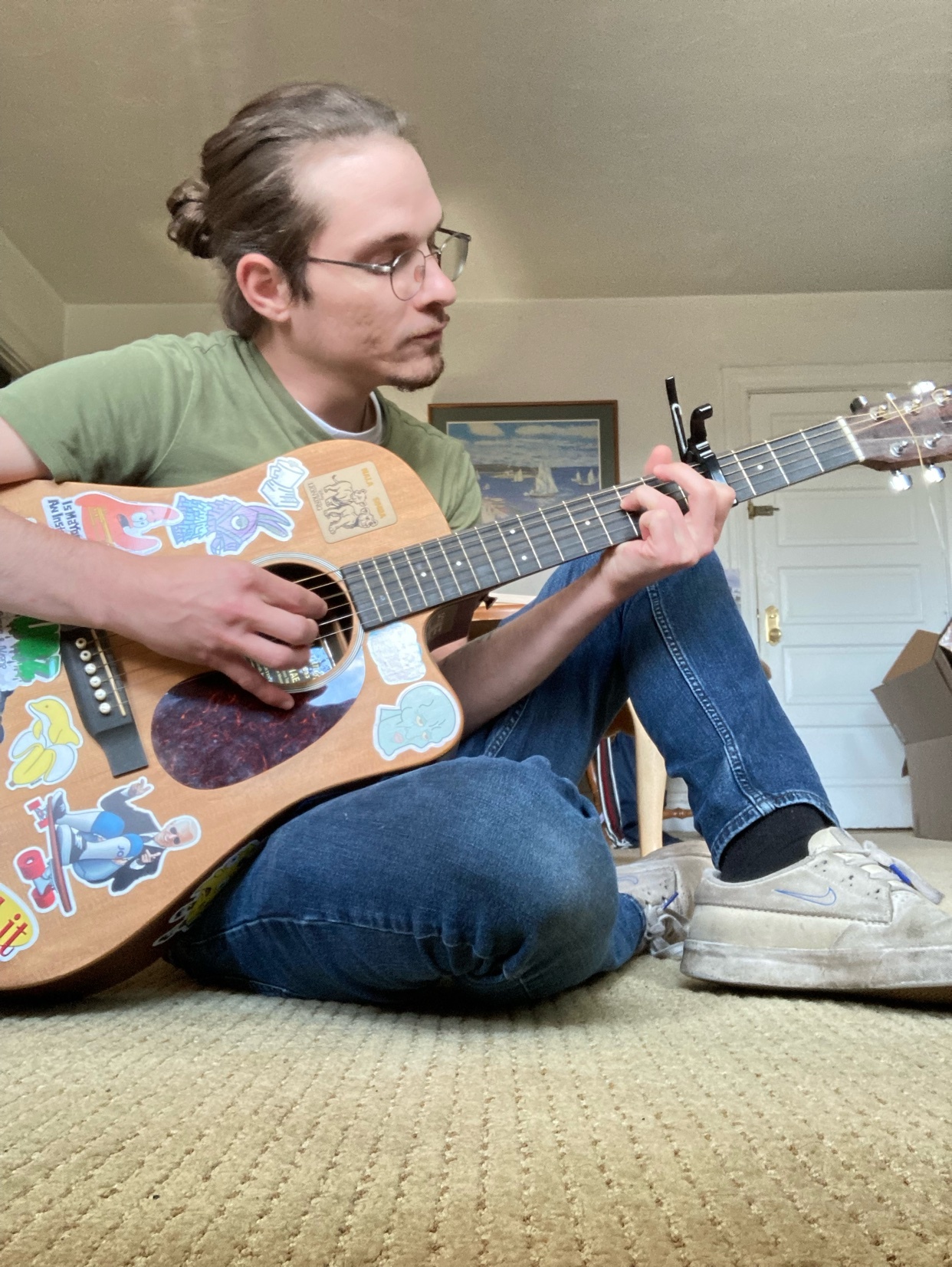 A man sits on the floor, playing an acoustic guitar. The guitar is covered in stickers, and he is wearing glasses.