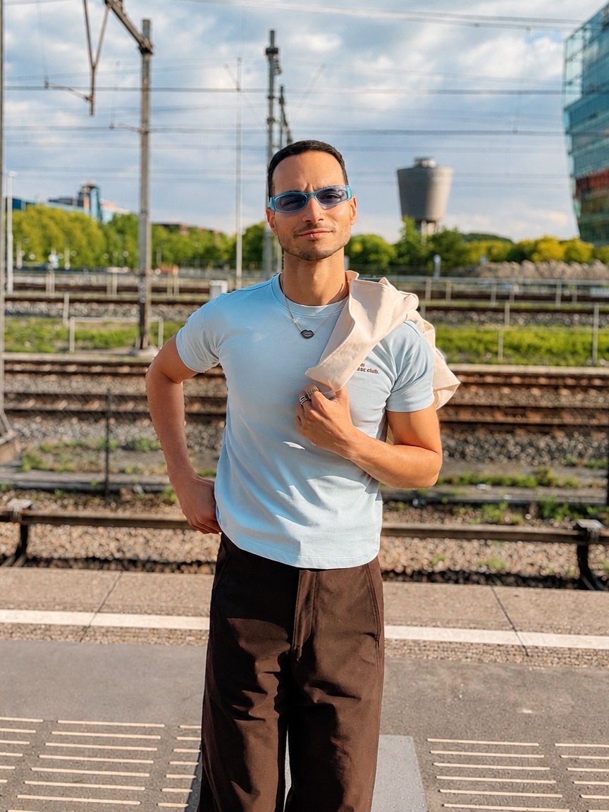 A man stands on a train platform, wearing sunglasses and a light blue t-shirt with brown pants. He has a light-colored sweater draped over his shoulder.