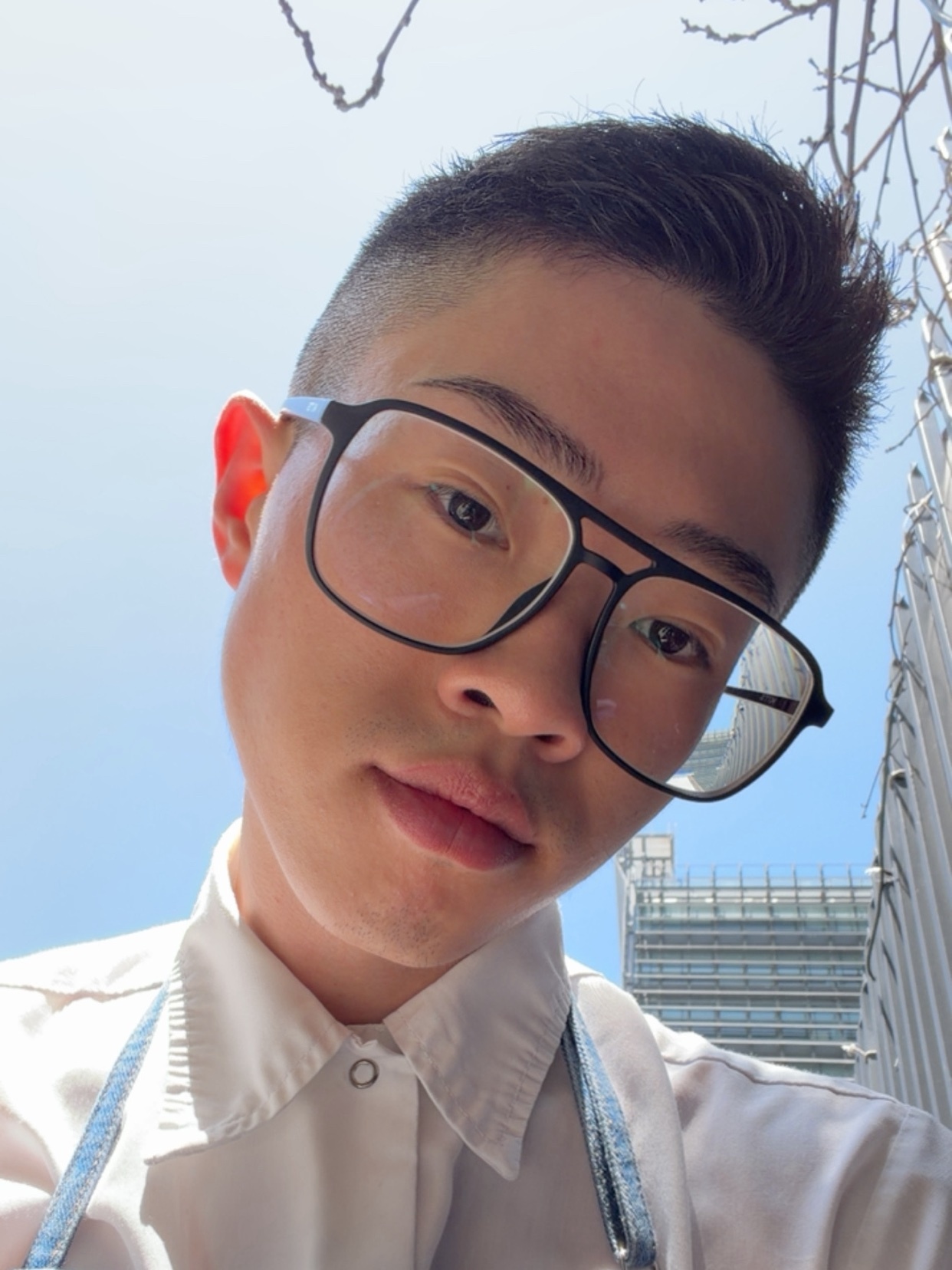 A close-up selfie of a young Asian man wearing black-rimmed glasses and a white button-up shirt. He is looking directly at the camera with a neutral expression.