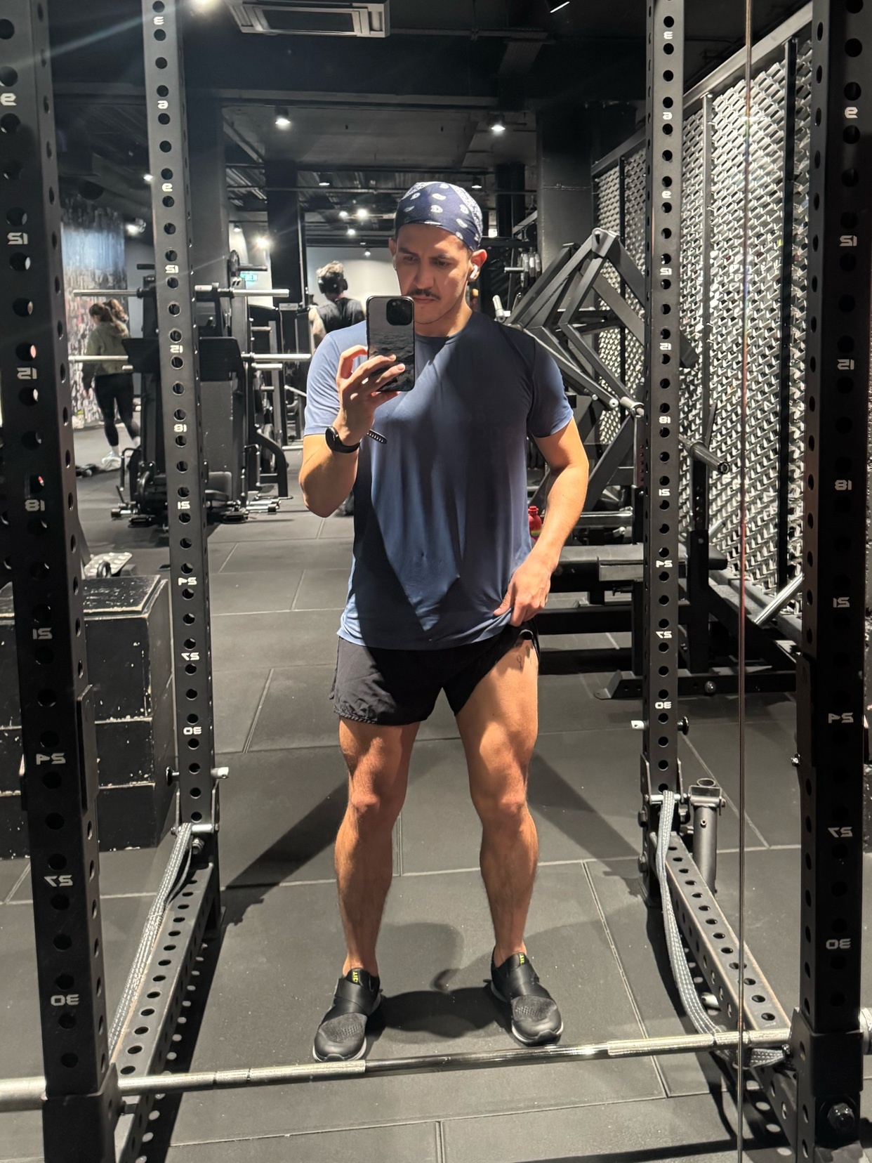 A man takes a mirror selfie at the gym, showcasing his muscular legs and physique. He's dressed in workout attire, ready for a fitness session.