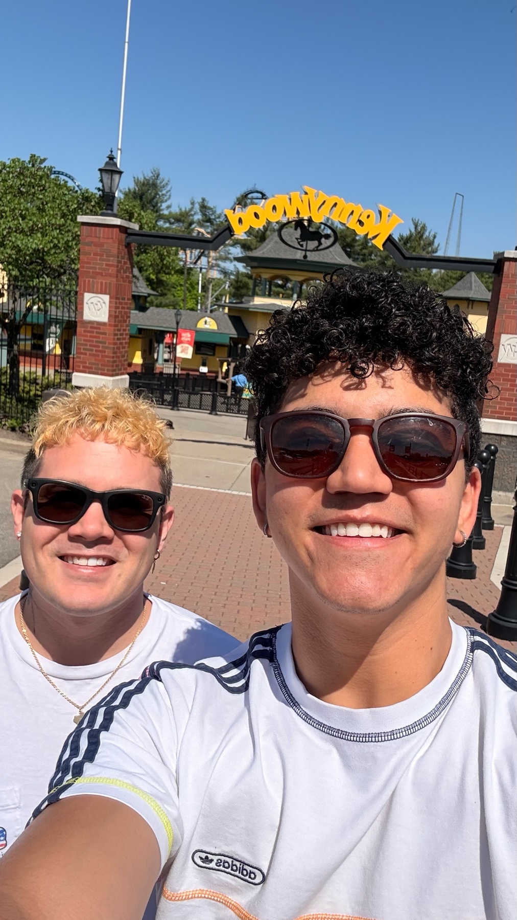 Two friends smile for a selfie in front of an amusement park entrance on a sunny day. Both are wearing sunglasses and casual t-shirts.