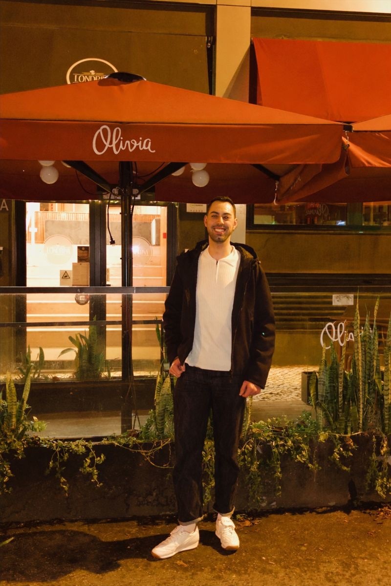 A man poses in front of a restaurant at night. He is smiling and wearing a casual outfit.