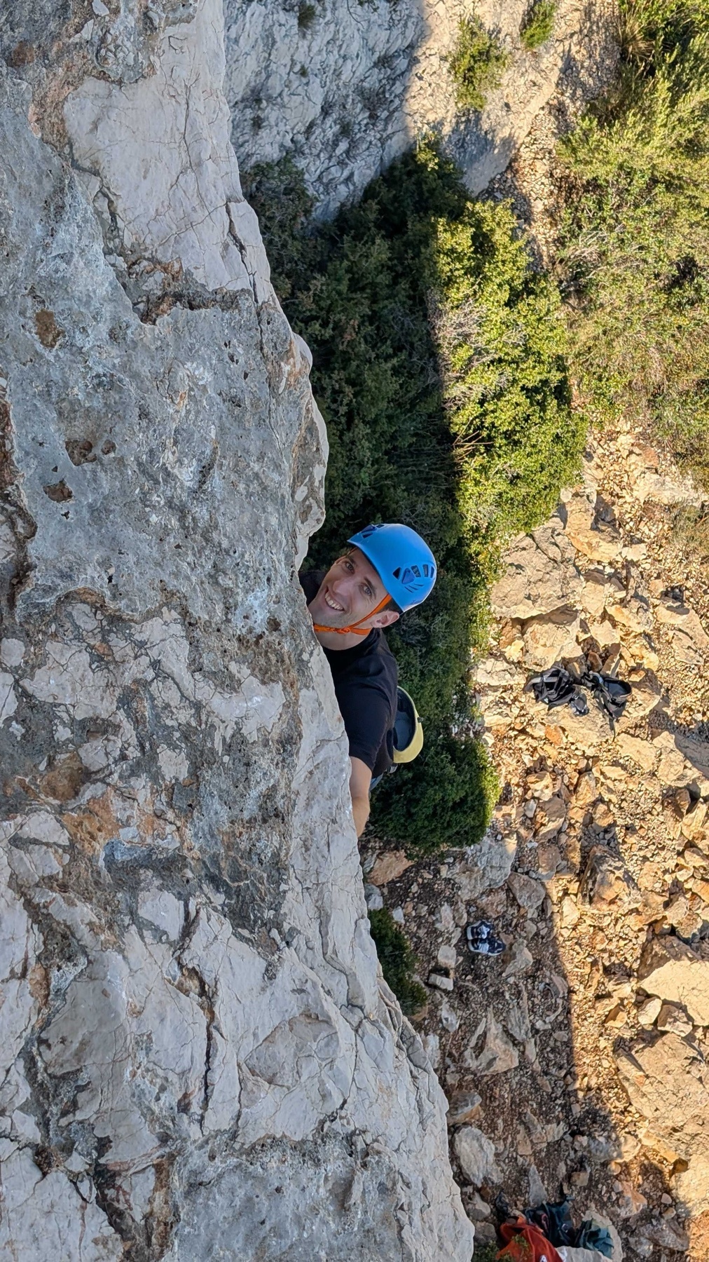 A man in a blue helmet and black t-shirt smiles as he climbs a rocky cliff face. He is wearing a yellow climbing harness and is surrounded by sparse vegetation and rocky terrain.
