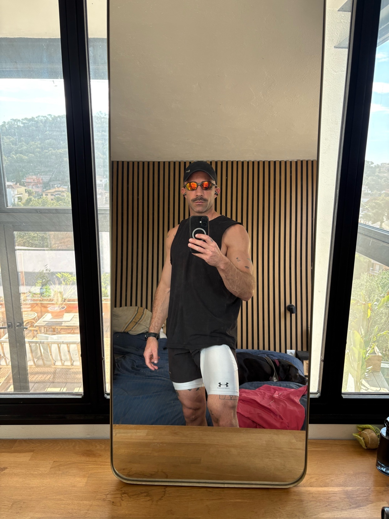 A man takes a mirror selfie wearing athletic wear, including a black tank top, white and black shorts, and sunglasses. He has a mustache and is wearing a baseball cap.