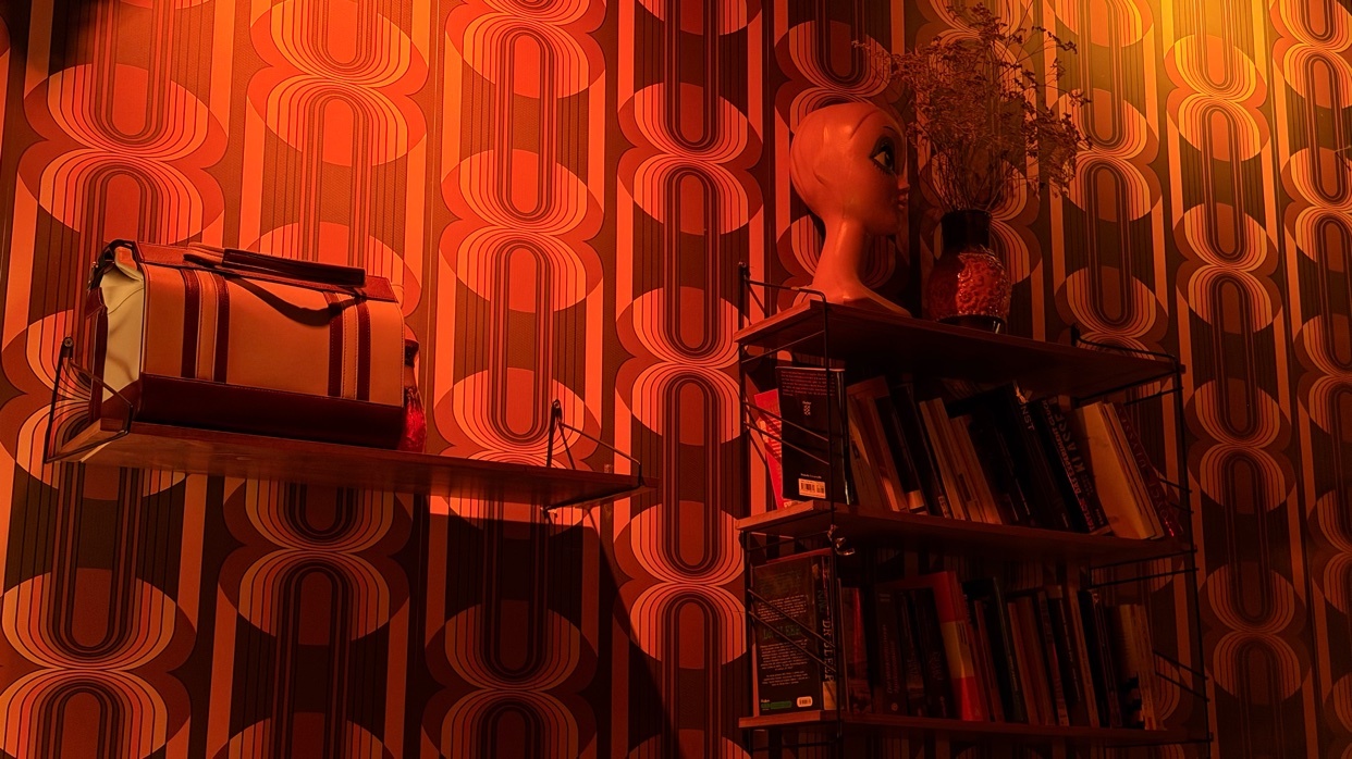 A vintage-style room features a bold, retro wallpaper with an orange and brown oval pattern. Two shelves are mounted on the wall, one holding a bag and the other displaying books and a mannequin head.