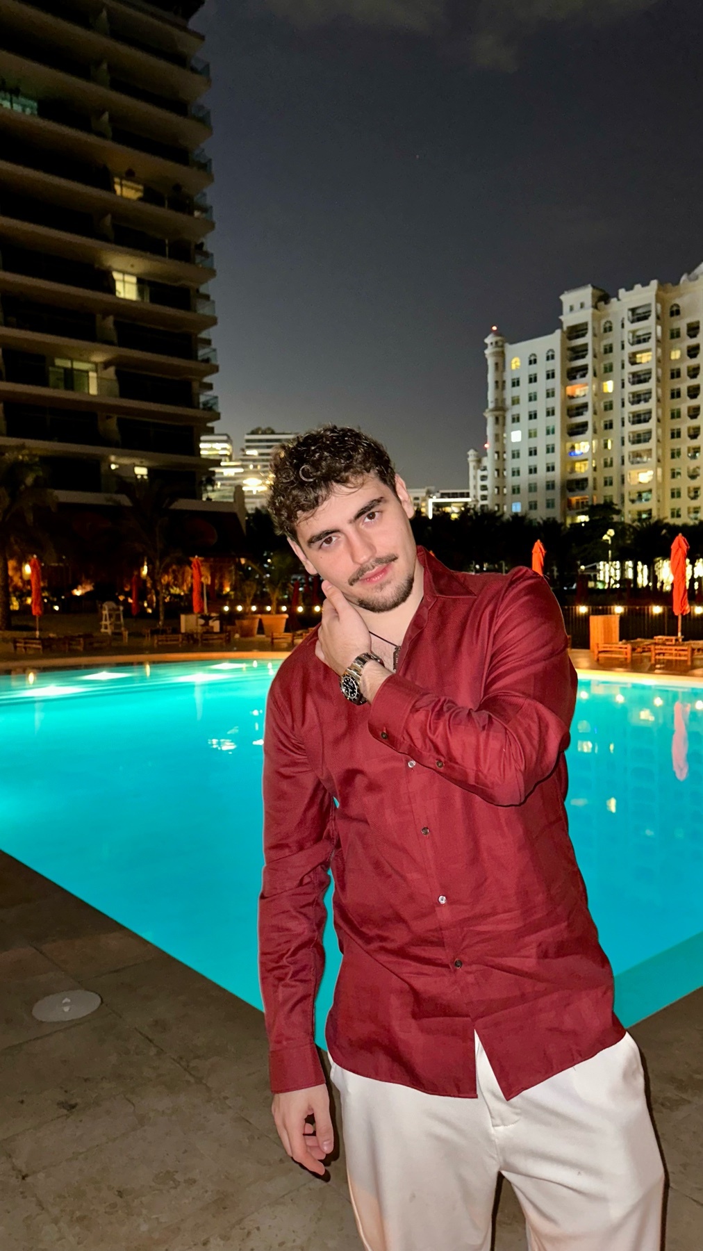 A man with curly hair and stubble poses by a swimming pool at night, with city buildings in the background.