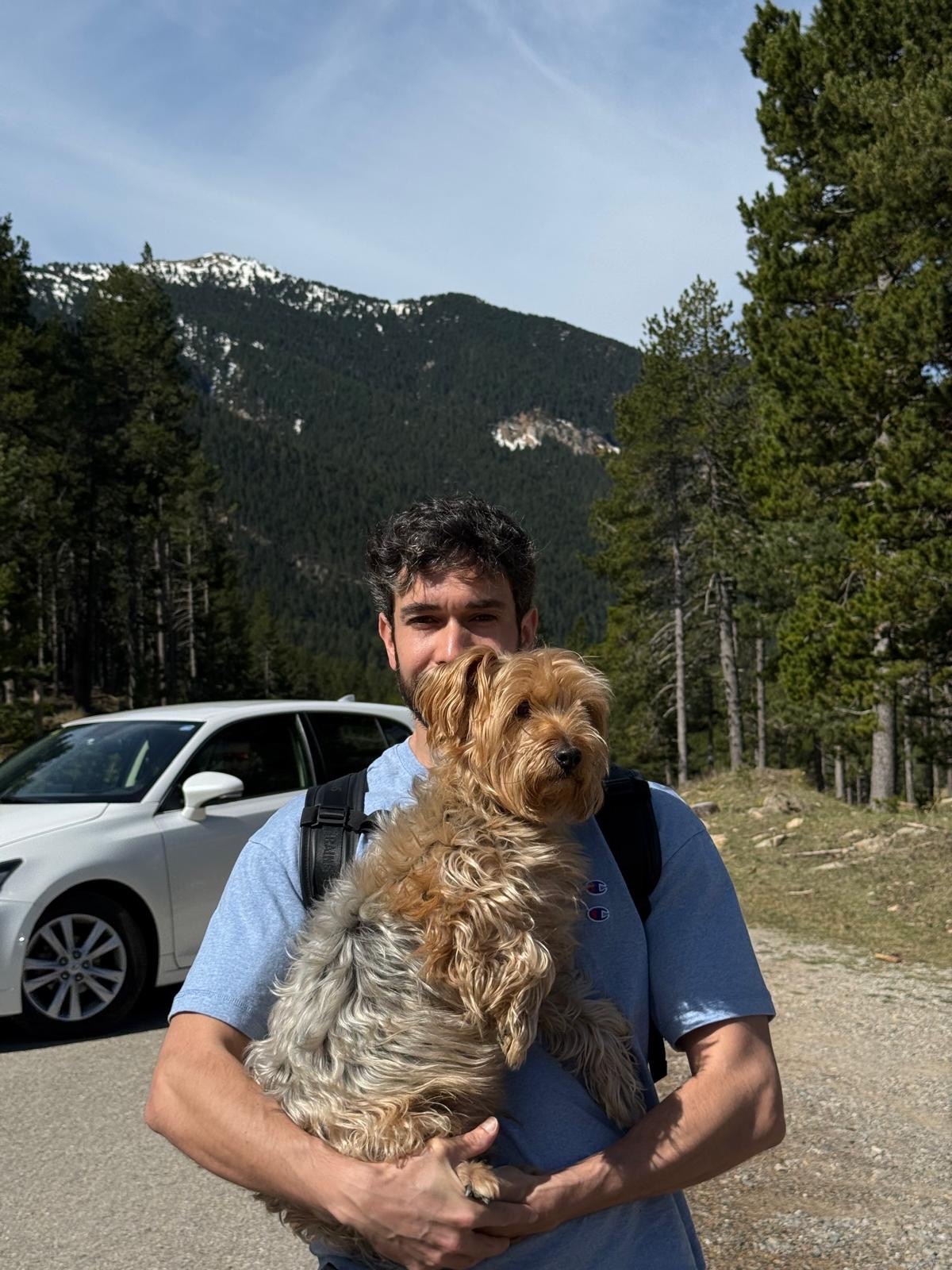 A man holds a small, tan dog in his arms while standing outdoors with a car and mountains in the background.