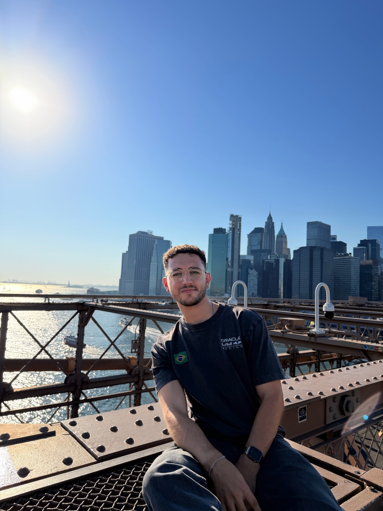 A man wearing glasses and a dark t-shirt poses for a photo on the Brooklyn Bridge with the New York City skyline in the background. The sun shines brightly in the clear blue sky.