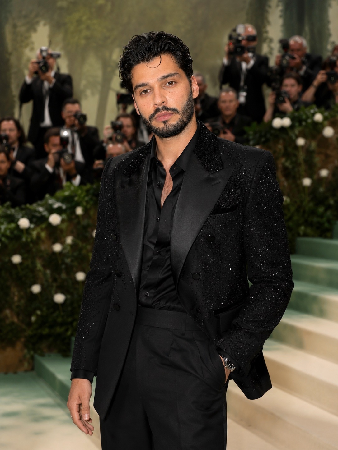 A man in a black suit with sparkling details poses for the camera at a formal event. He has dark, curly hair, stubble, and a beard, and is wearing a black button-up shirt underneath his suit jacket.