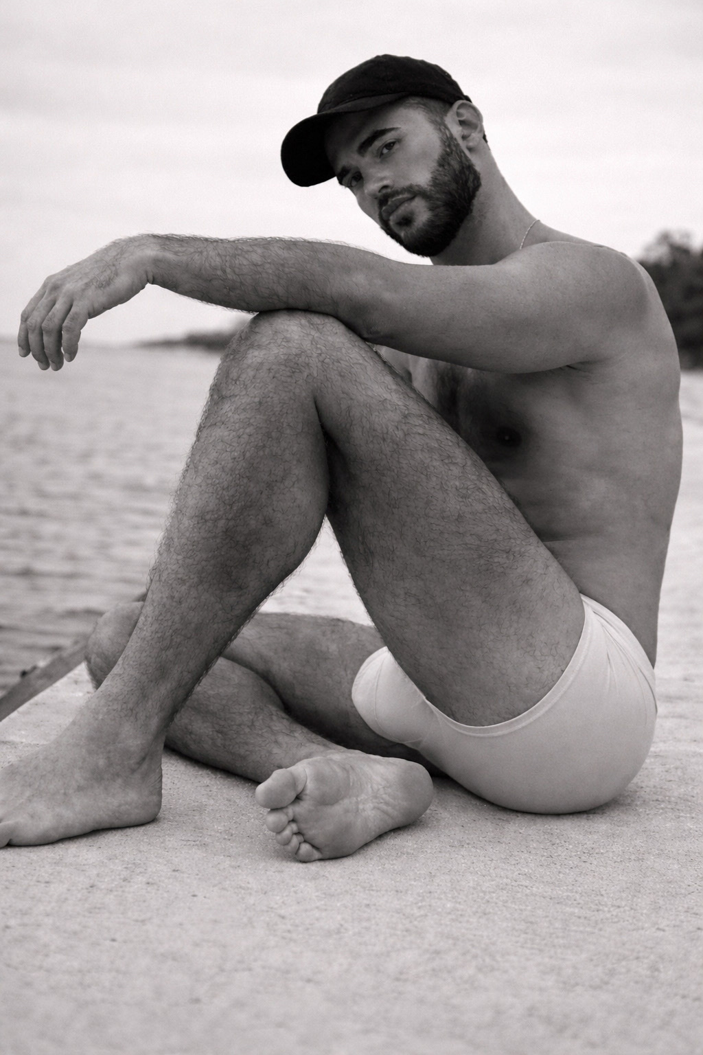 A black and white photo of a man sitting shirtless on a dock. He is wearing a cap and underwear.