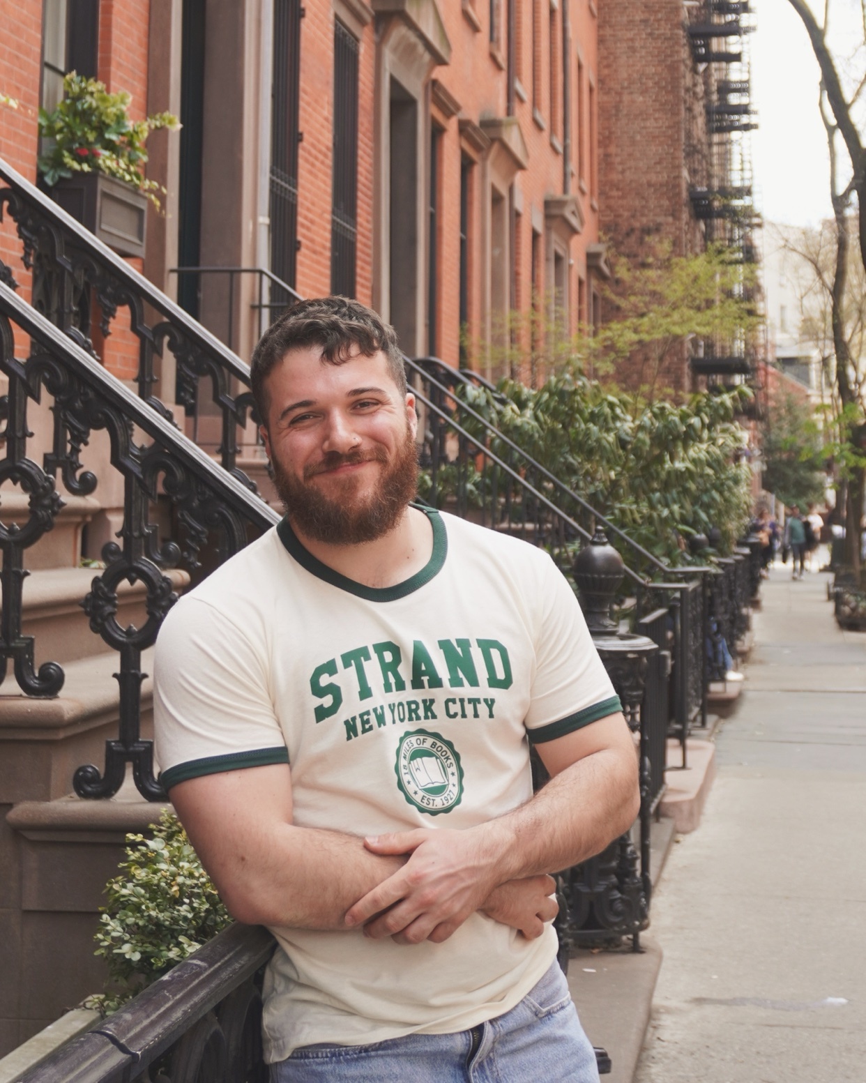 A man with a beard smiles at the camera, leaning against a railing in front of a brick building in New York City. He is wearing a t-shirt that says "STRAND NEW YORK CITY" and jeans.