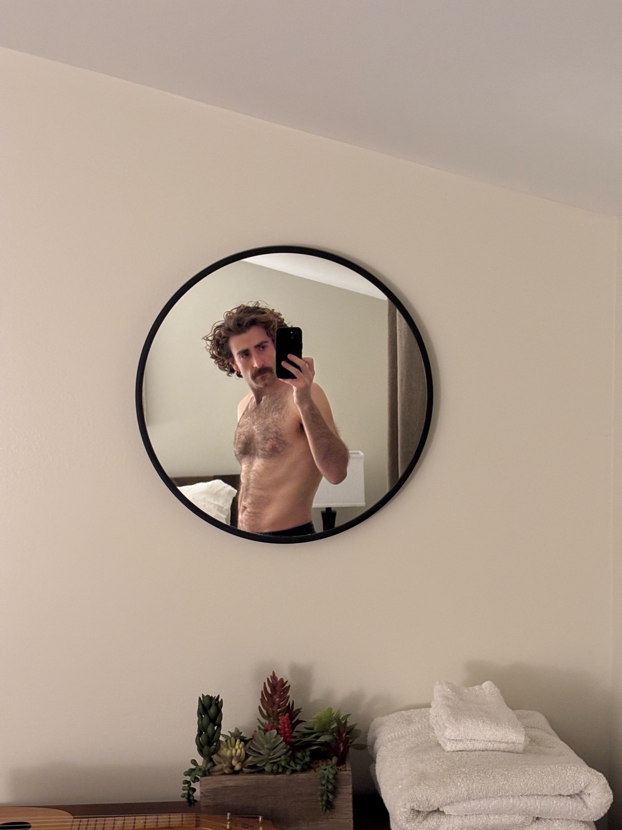 A man takes a shirtless selfie in a round mirror. The photo is taken in a bedroom with a modern aesthetic.