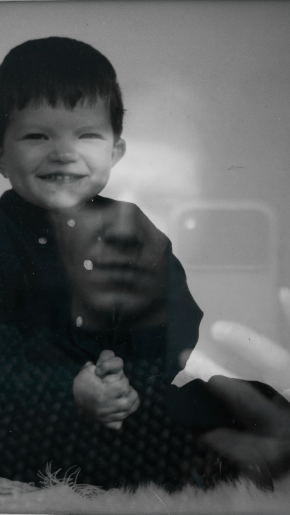 A black and white photo shows a smiling child in the foreground, with a faint overlay of an adult's face behind them. The image has a vintage, nostalgic feel.