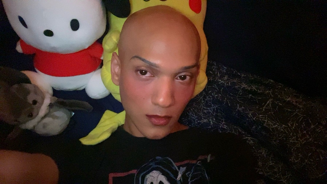 A person with a bald head and makeup poses for a selfie, surrounded by plush toys including Hello Kitty and Pikachu.