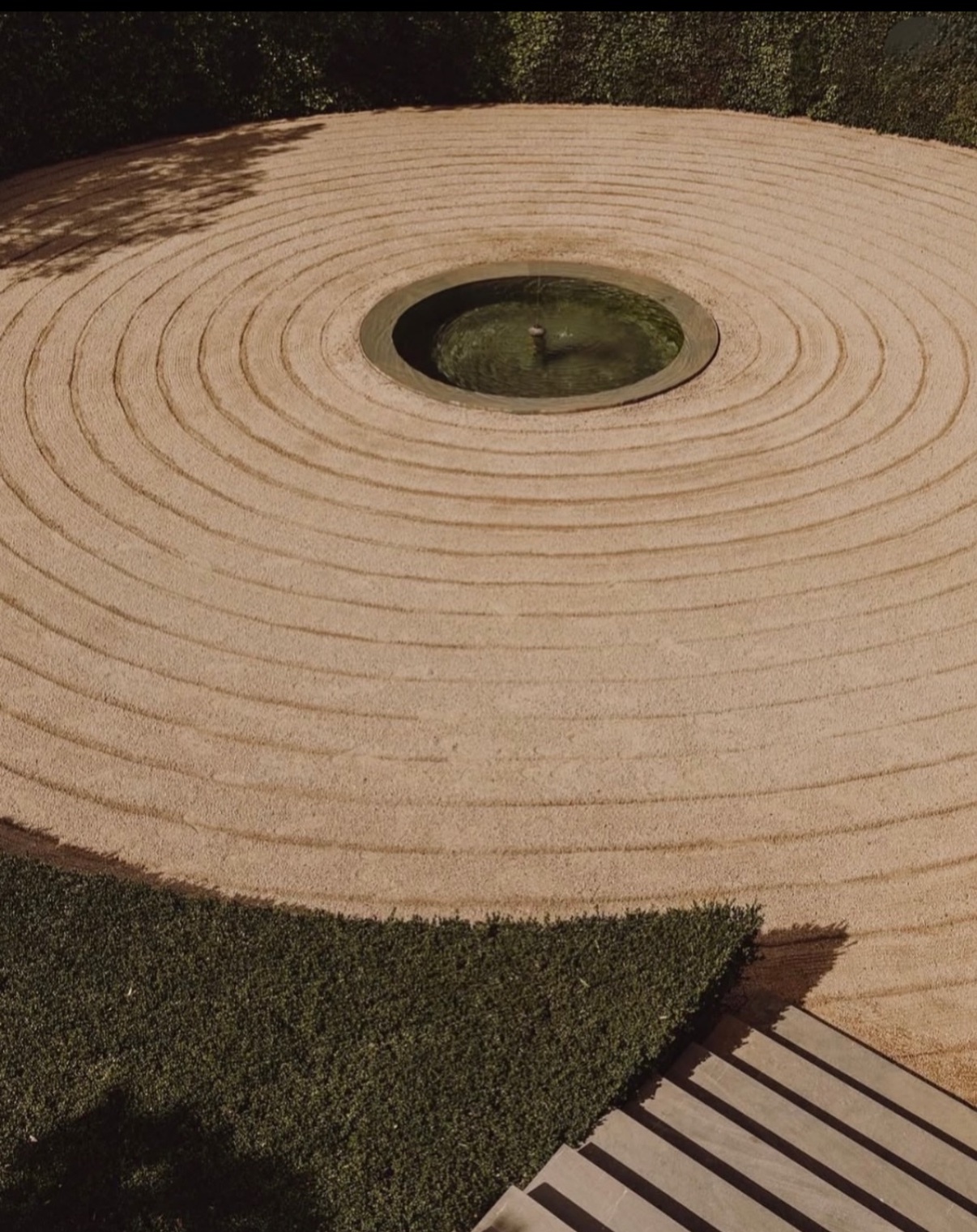 A serene zen garden features a circular pattern raked into the sand, surrounding a central water feature. Lush green grass borders the sand, with a hint of modern landscaping visible.