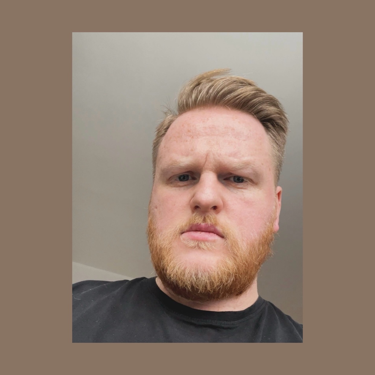 A man with blonde hair and a red beard looks directly at the camera. He is wearing a black t-shirt.