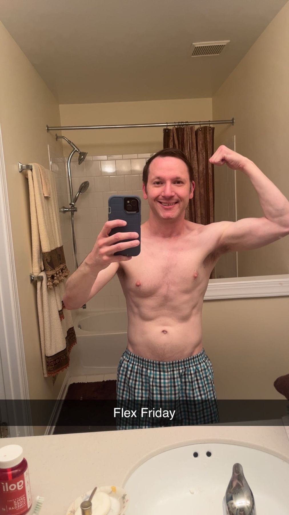 A man in plaid boxer shorts poses in a bathroom mirror, flexing his bicep and showing off his abs. The text 'Flex Friday' is overlaid on the image.
