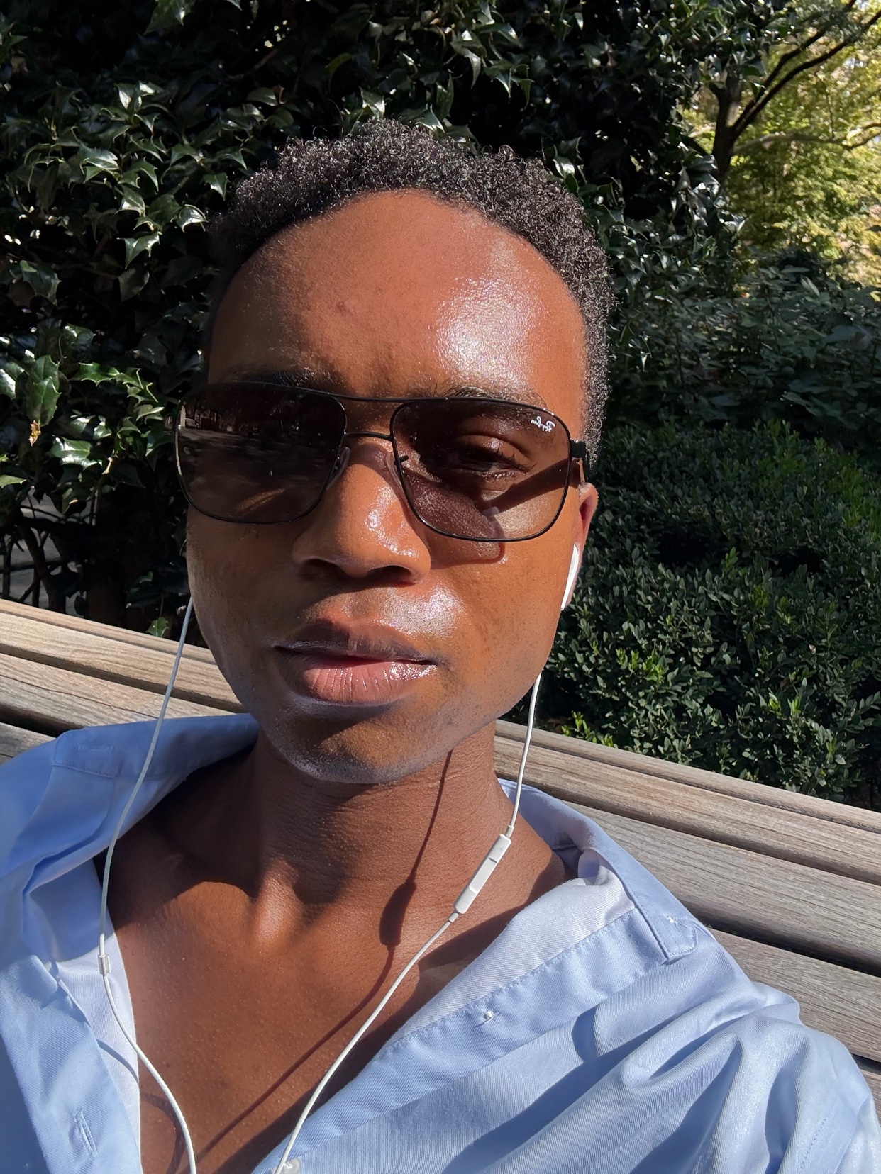 A man wearing sunglasses and earbuds takes a selfie outdoors on a sunny day. He is wearing a light blue collared shirt and sitting on a wooden bench.