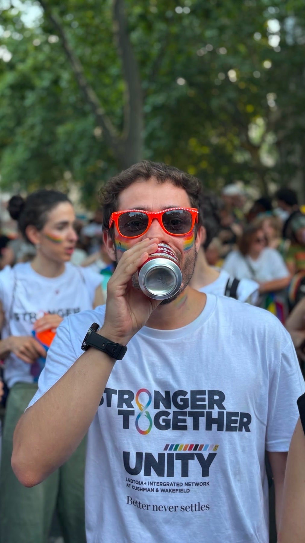 A man wearing bright orange sunglasses and a white t-shirt with the words "STRONGER TOGETHER UNITY" printed on it, drinks from a can. He is in a crowd of people, suggesting a festive event.