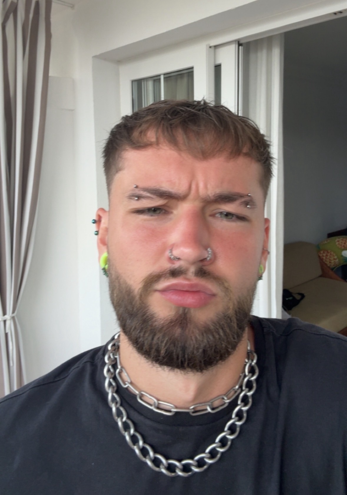 A man with multiple facial piercings and a chain necklace poses for a selfie. He has dark hair and a beard.