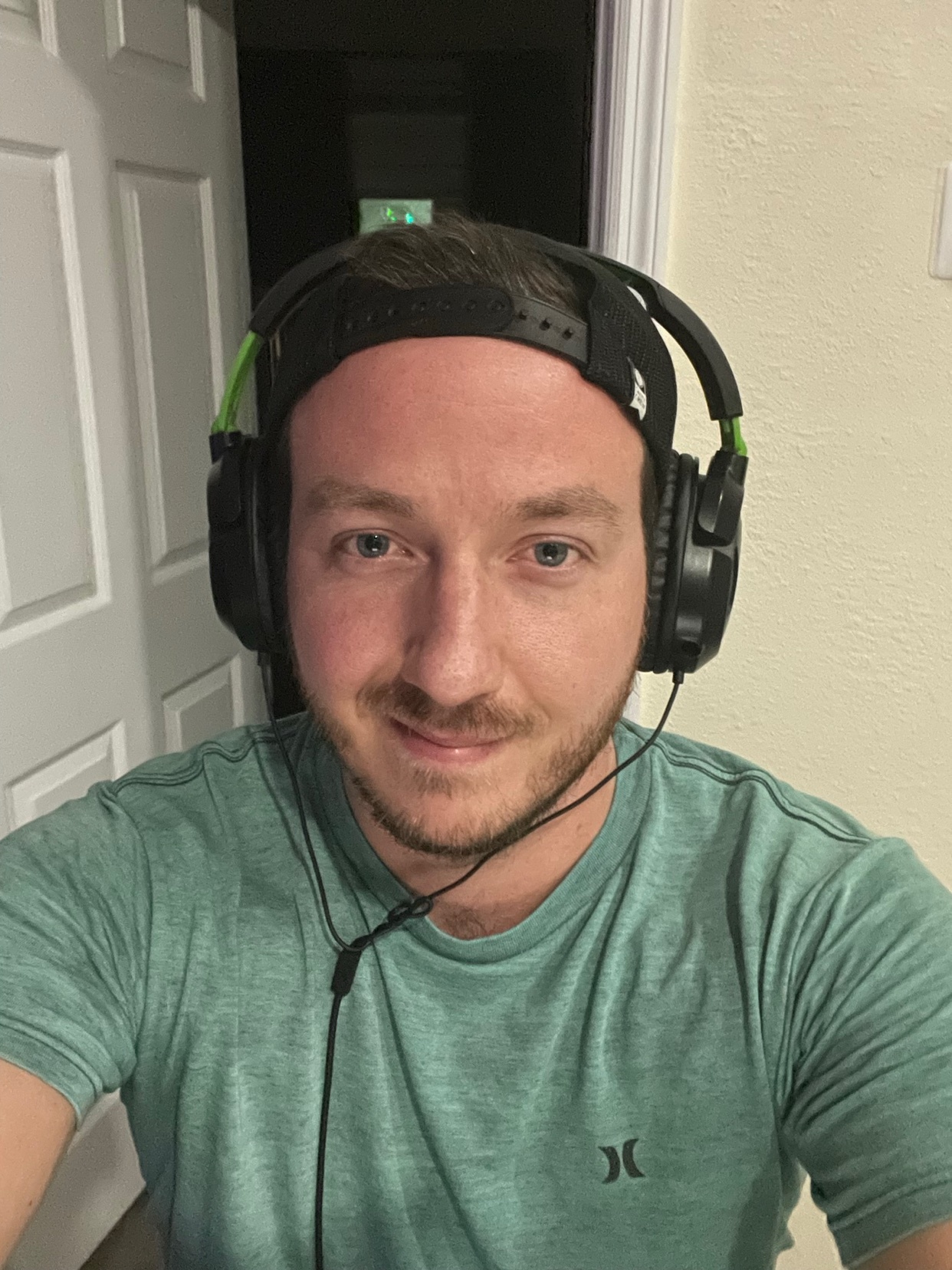 A man with stubble and a beard smiles at the camera while wearing a green t-shirt and a black gaming headset with green accents.