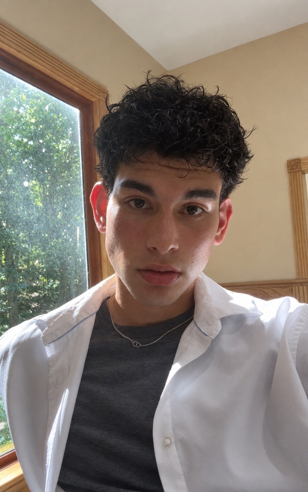 A young man with curly dark hair poses for a selfie indoors, wearing a grey t-shirt and an unbuttoned white shirt. Sunlight streams in from a window behind him.