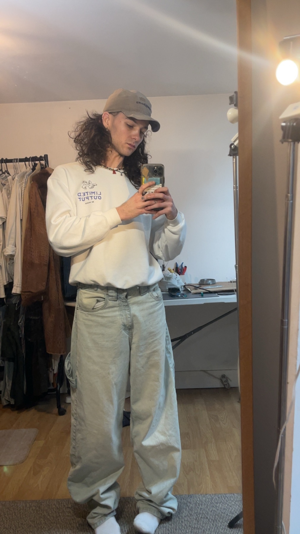 A man takes a mirror selfie, showcasing his outfit. He wears a cap, sweatshirt, and baggy jeans.