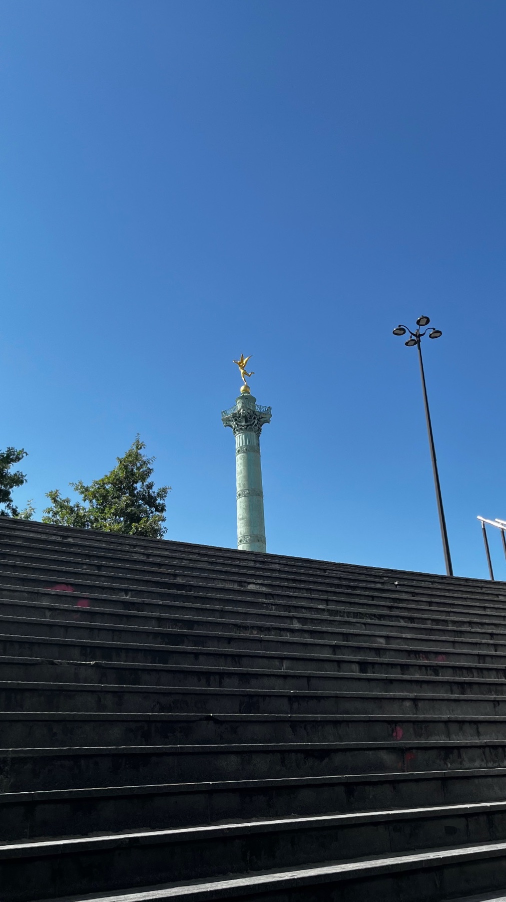A tall column monument topped with a golden statue stands against a clear blue sky, viewed from the bottom of a wide set of outdoor stairs.