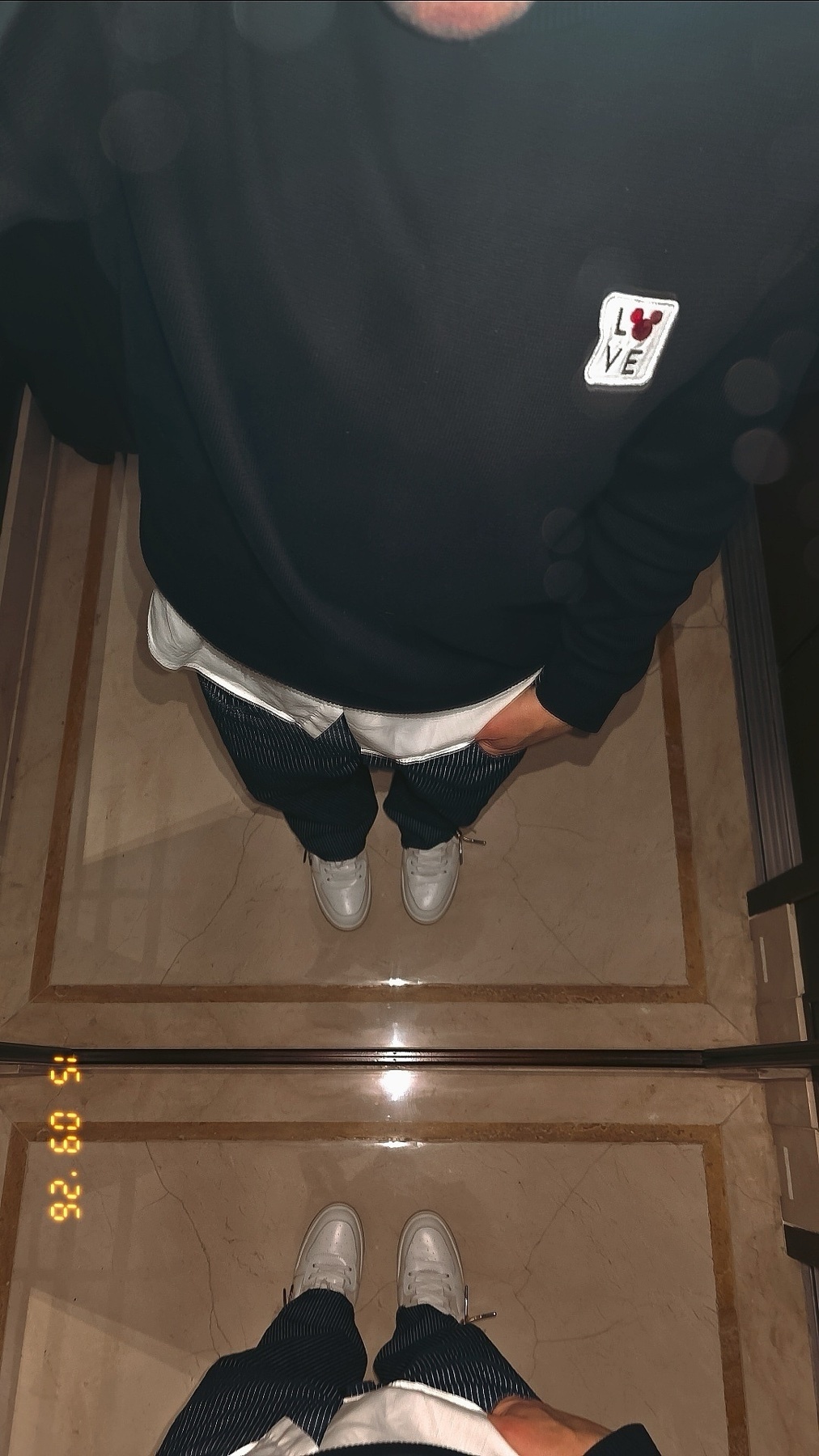 A couple takes a mirror selfie, showcasing their matching white sneakers and casual outfits. The reflection captures their legs and feet against a marble floor.