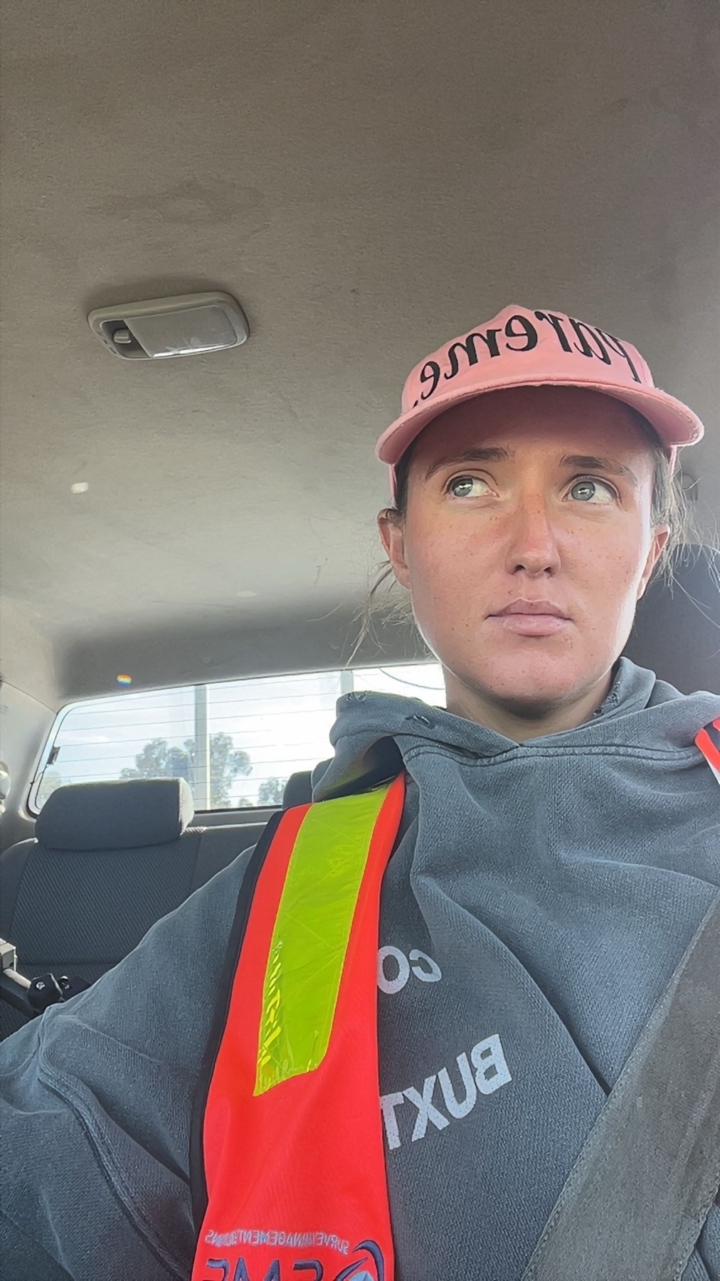 A woman wearing a pink baseball cap and a grey hoodie with a reflective safety vest over it takes a selfie inside a car.