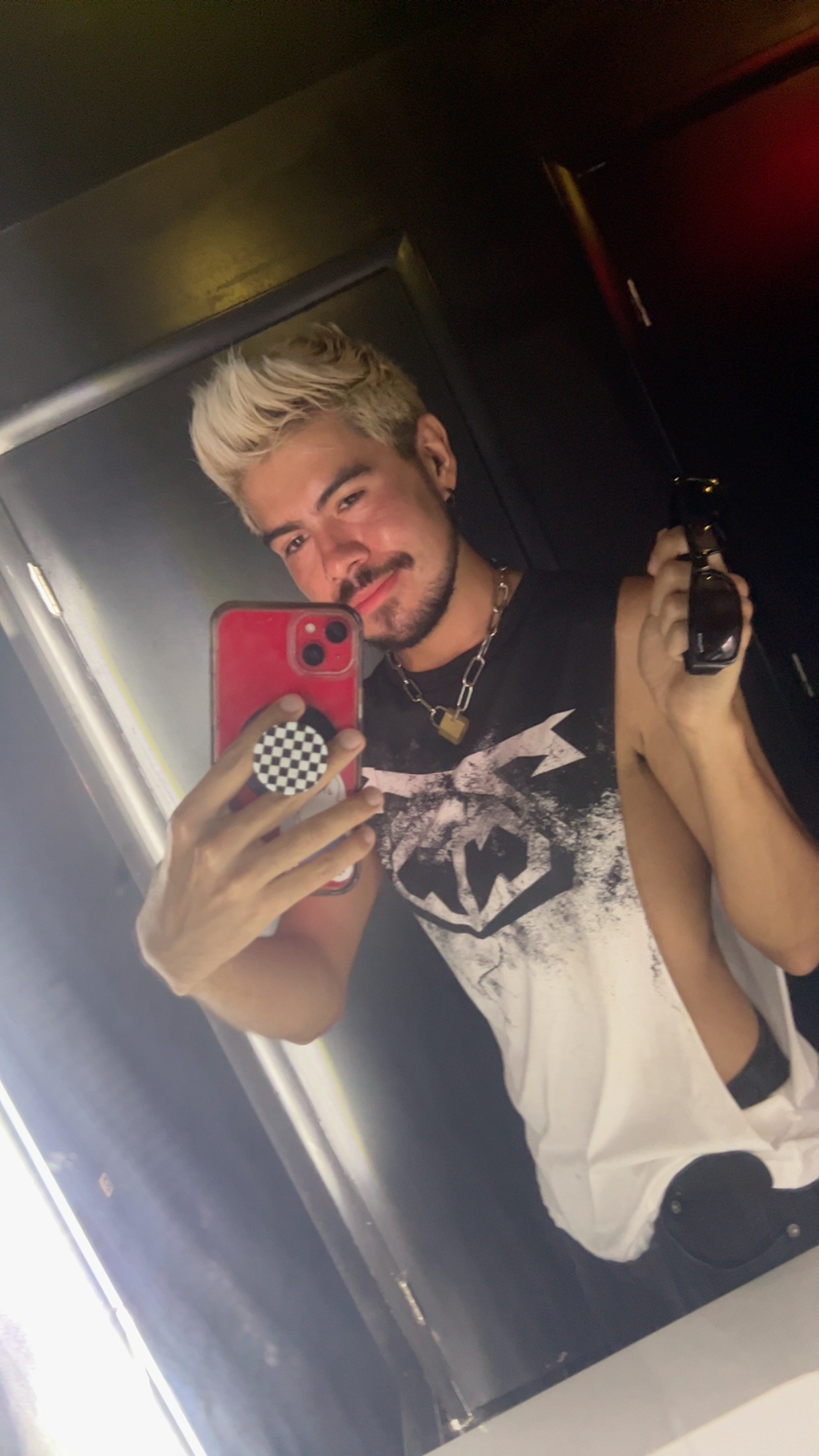 A man with blonde hair and stubble takes a selfie in a dimly lit room, wearing a graphic tank top and a chain necklace.