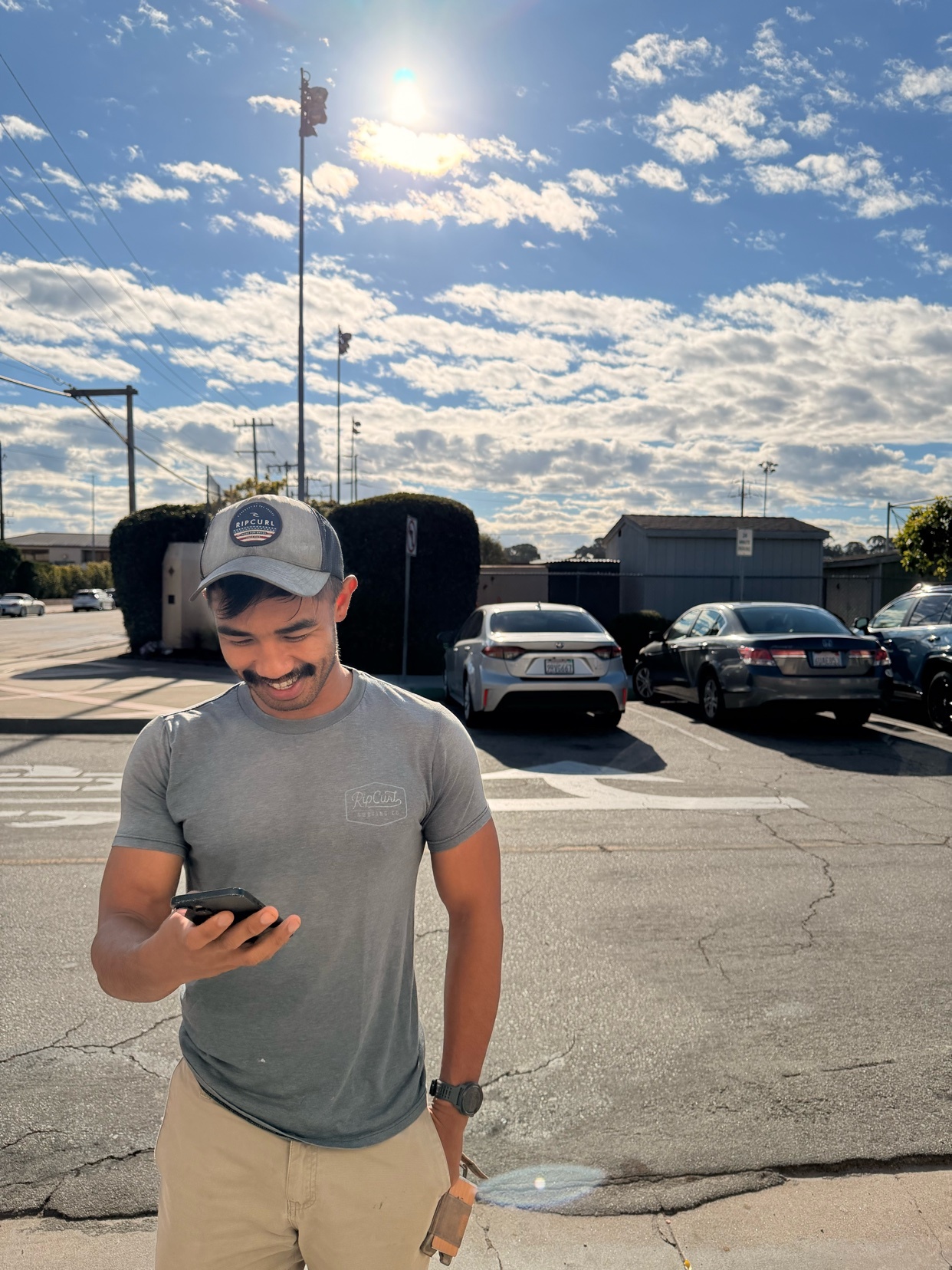 A man with a mustache and a hat looks down at his phone while standing in a parking lot under a bright, cloudy sky. Cars are parked in the background.