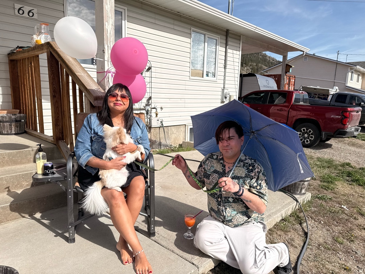 A woman and a man are sitting outside a house with a dog and balloons. The woman is holding a small white dog while sitting in a chair. The man is sitting on the ground holding a blue umbrella. There are two balloons, one pink and one white, tied to